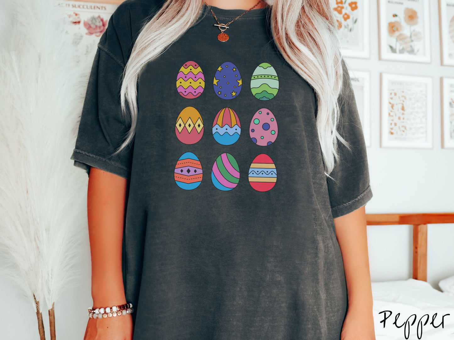 A woman wearing a cute, vintage pepper colored shirt with a three by three grid of colorful Easter eggs. One is pink, red, and yellow, another blue with stars, and another is different shades of green next to five more egg designs.
