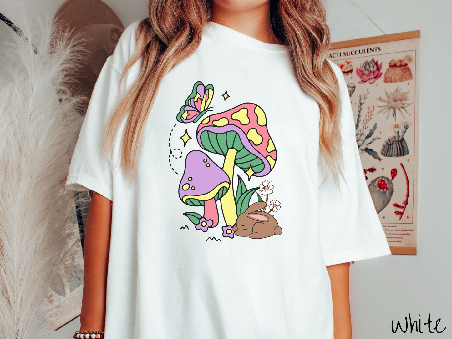 A woman wearing a cute, vintage white colored shirt with a purple mushroom and a pink and yellow mushroom together with a brown bunny rabbit laying on the ground by purple and white flowers, and a colorful butterfly is fluttering above it all.