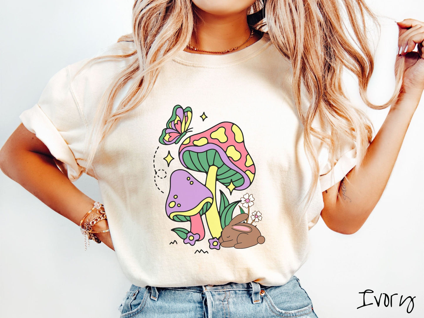 A woman wearing a cute, vintage ivory colored shirt with a purple mushroom and a pink and yellow mushroom together with a brown bunny rabbit laying on the ground by purple and white flowers, and a colorful butterfly is fluttering above it all.