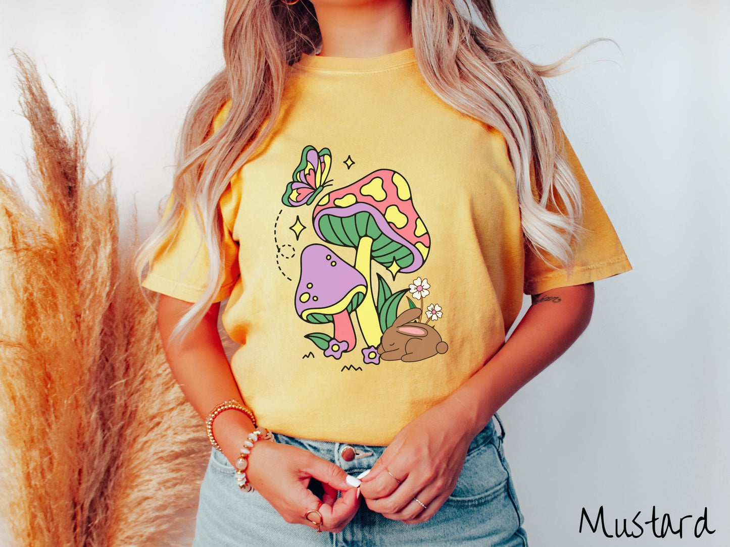 A woman wearing a cute, vintage mustard colored shirt with a purple mushroom and a pink and yellow mushroom together with a brown bunny rabbit laying on the ground by purple and white flowers, and a colorful butterfly is fluttering above it all.