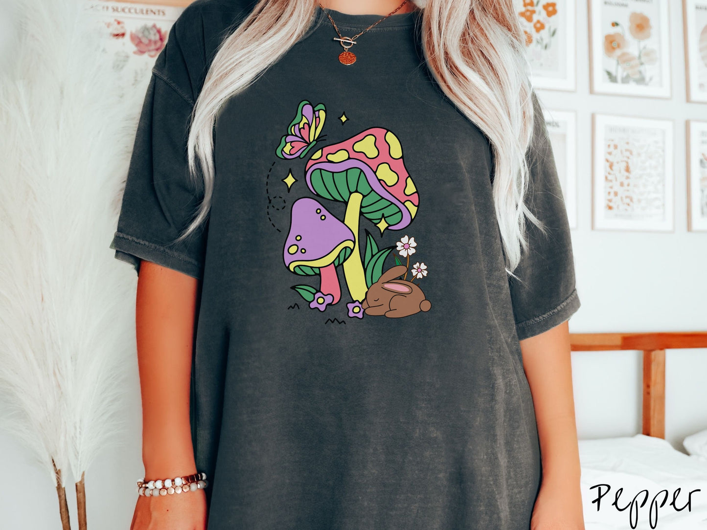 A woman wearing a cute, vintage pepper colored shirt with a purple mushroom and a pink and yellow mushroom together with a brown bunny rabbit laying on the ground by purple and white flowers, and a colorful butterfly is fluttering above it all.