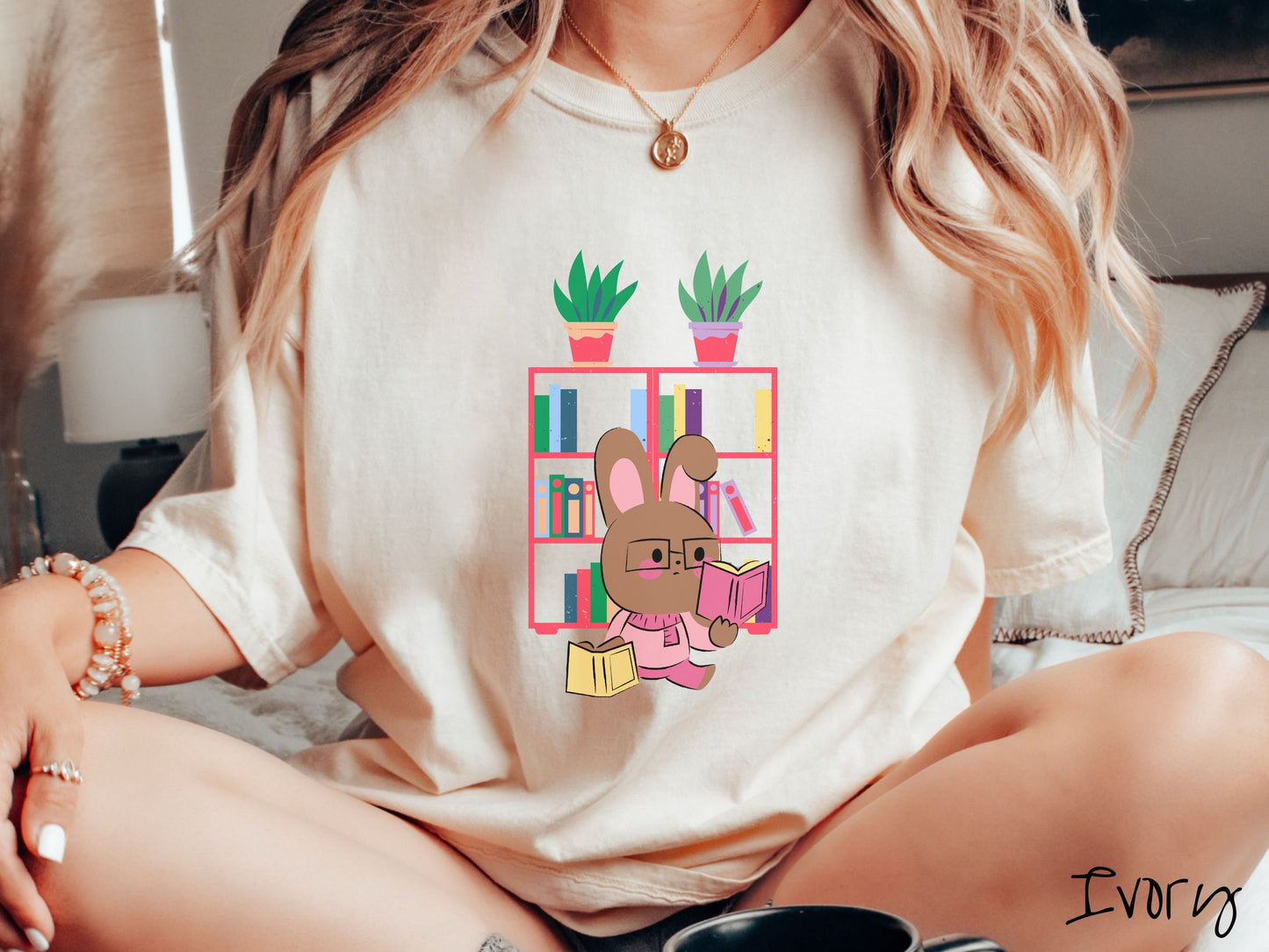 A woman wearing a cute, vintage ivory colored shirt with a pink-cheeked, brown bunny rabbit wearing glasses reading a pink book in one hand, and a yellow book in the other. In the background is a bookshelf with green plants on top.