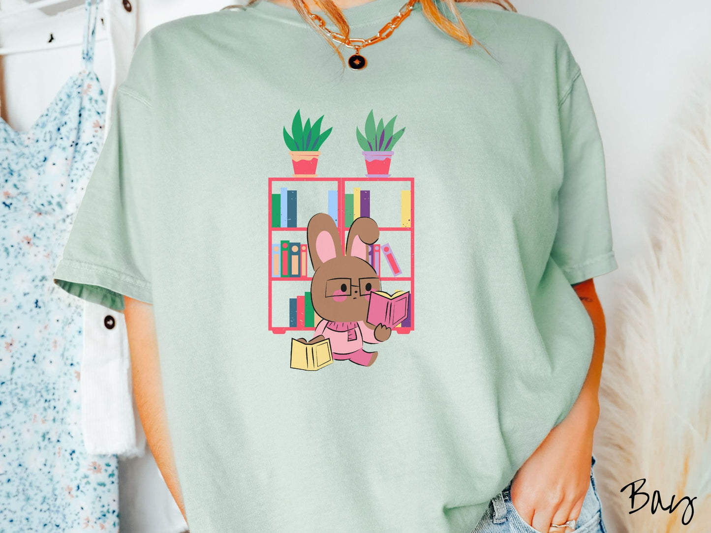 A woman wearing a cute, vintage bay colored shirt with a pink-cheeked, brown bunny rabbit wearing glasses reading a pink book in one hand, and a yellow book in the other. In the background is a bookshelf with green plants on top.