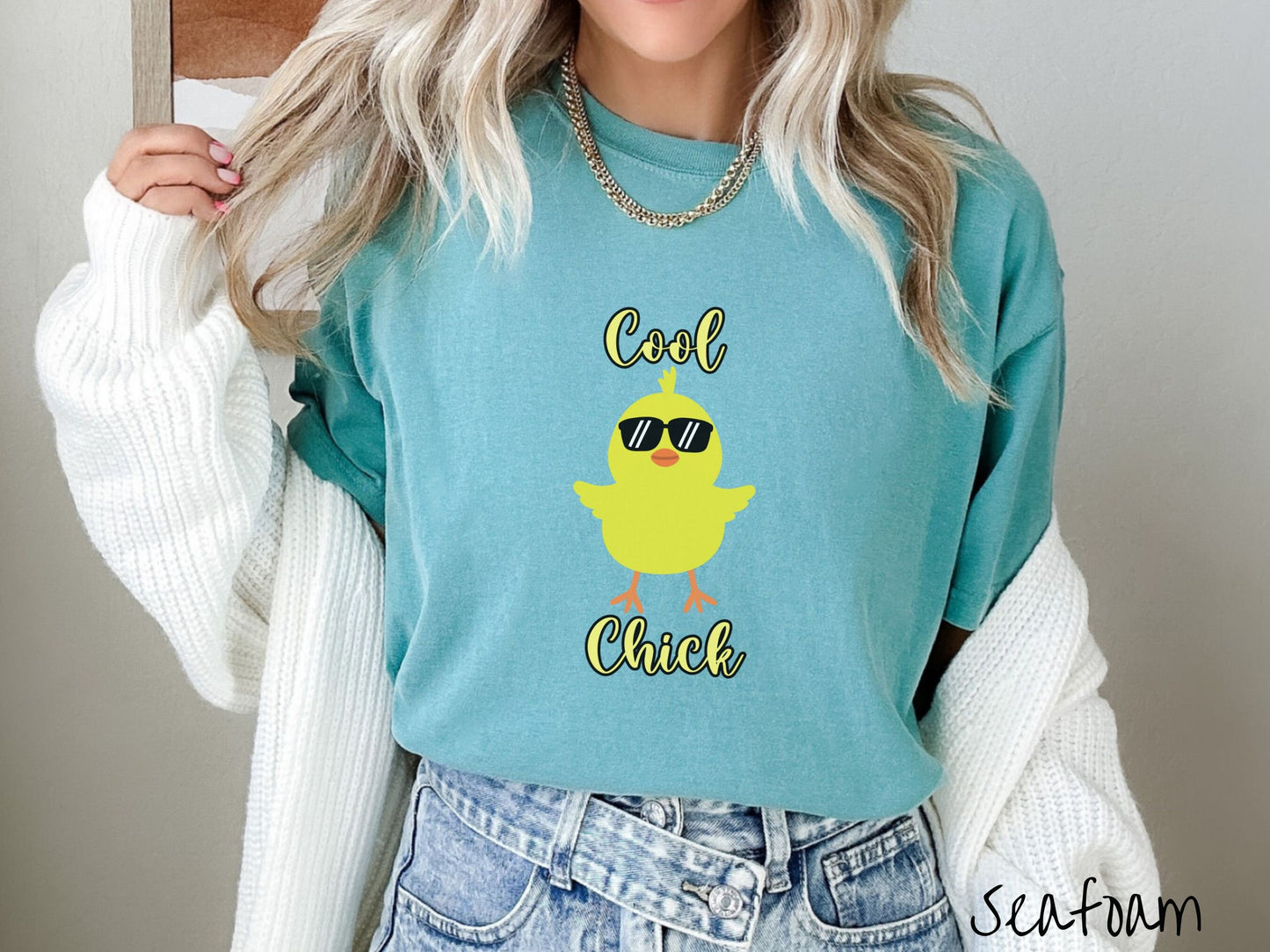 A woman wearing a cute, vintage seafoam colored shirt with the yellow text Cool Chick and below is a yellow baby chick with black sunglasses with its wings spread. It has an orange mouth and feet and yellow feathers sticking out the top of its head.