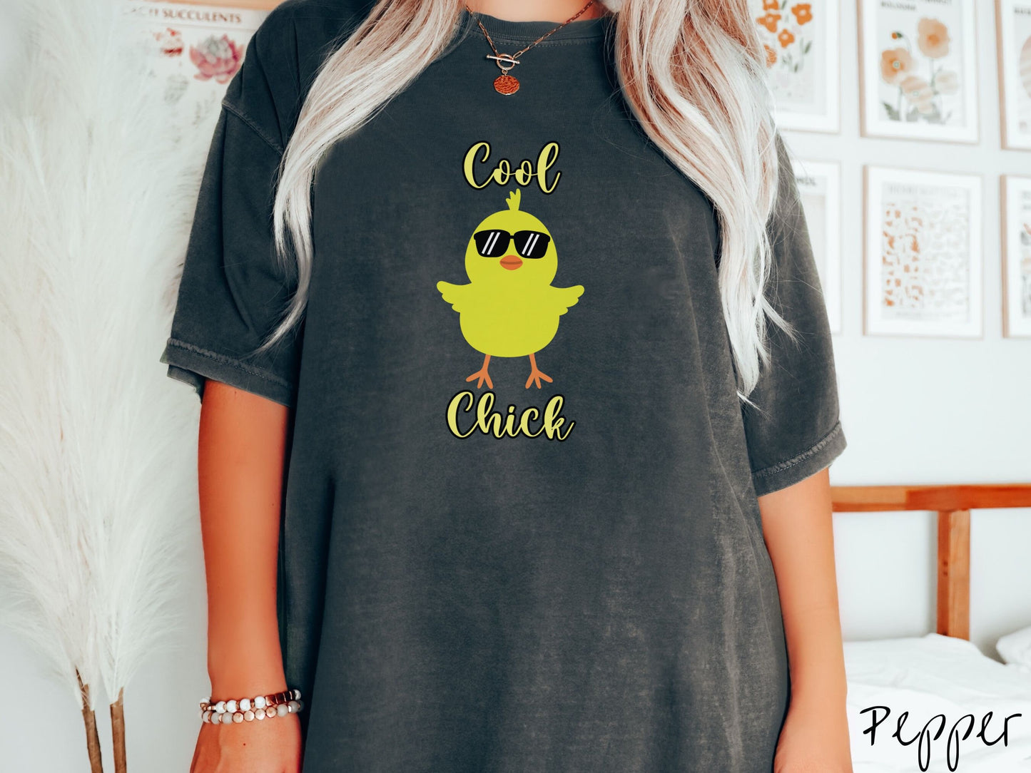 A woman wearing a cute, vintage pepper colored shirt with the yellow text Cool Chick and below is a yellow baby chick with black sunglasses with its wings spread. It has an orange mouth and feet and yellow feathers sticking out the top of its head.