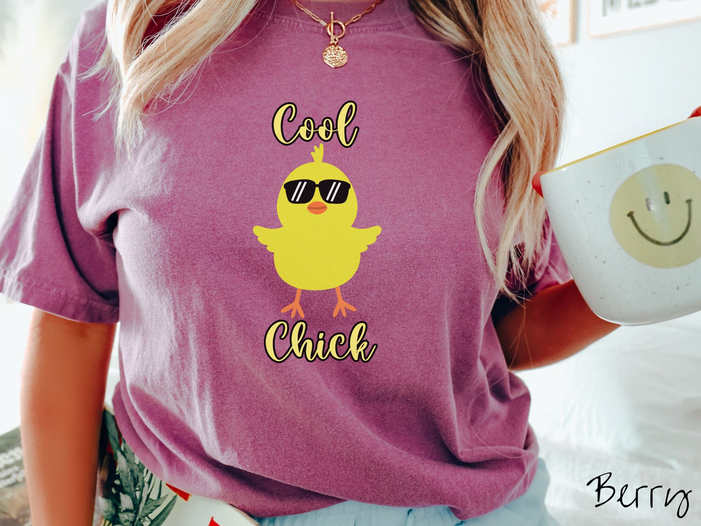 A woman wearing a cute, vintage berry colored shirt with the yellow text Cool Chick and below is a yellow baby chick with black sunglasses with its wings spread. It has an orange mouth and feet and yellow feathers sticking out the top of its head.