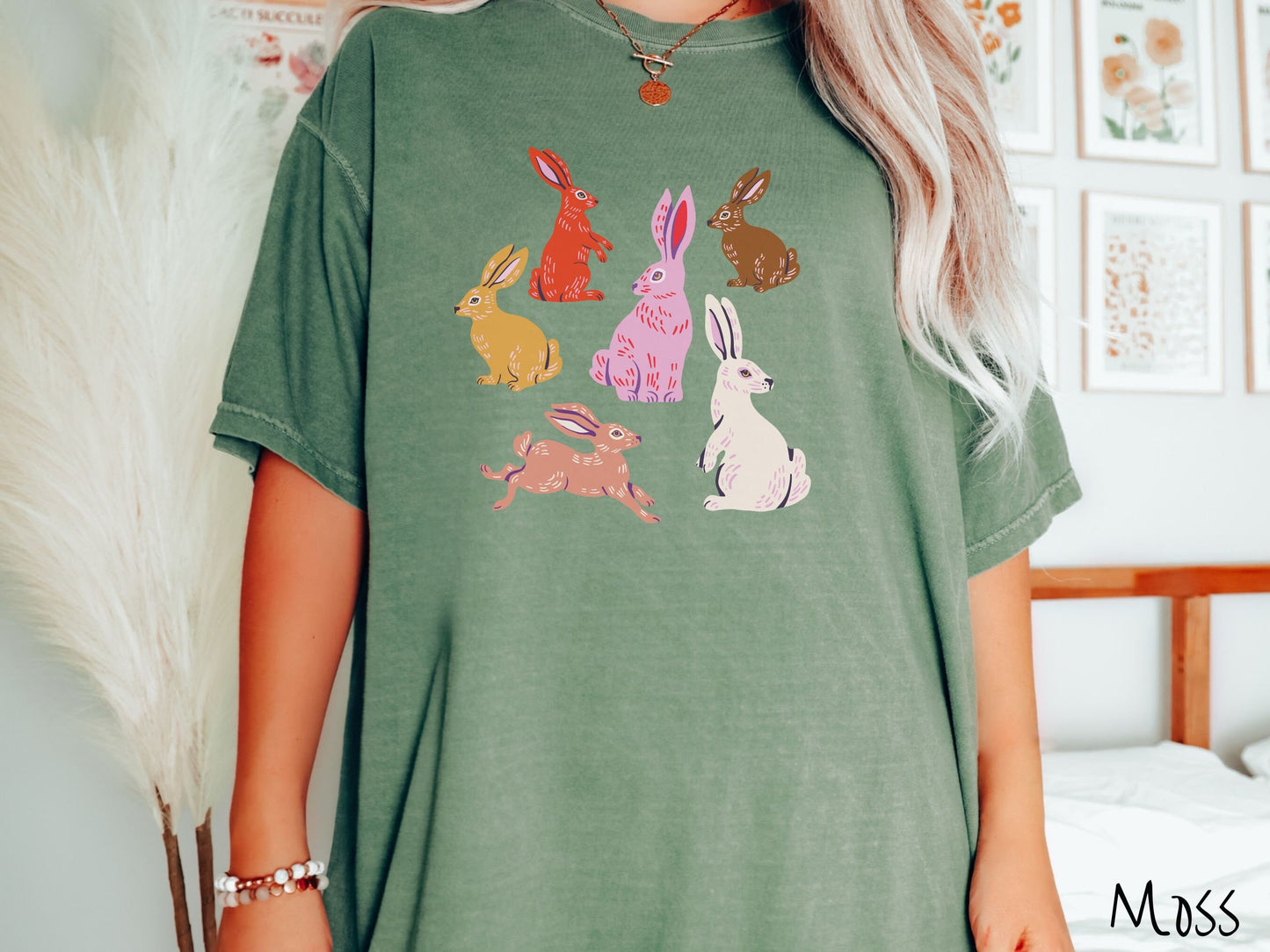 A woman wearing a cute, vintage moss colored shirt with six colored bunny rabbits in different stances and postures. One is red, another brown, yellow, pink, light brown, and white.
