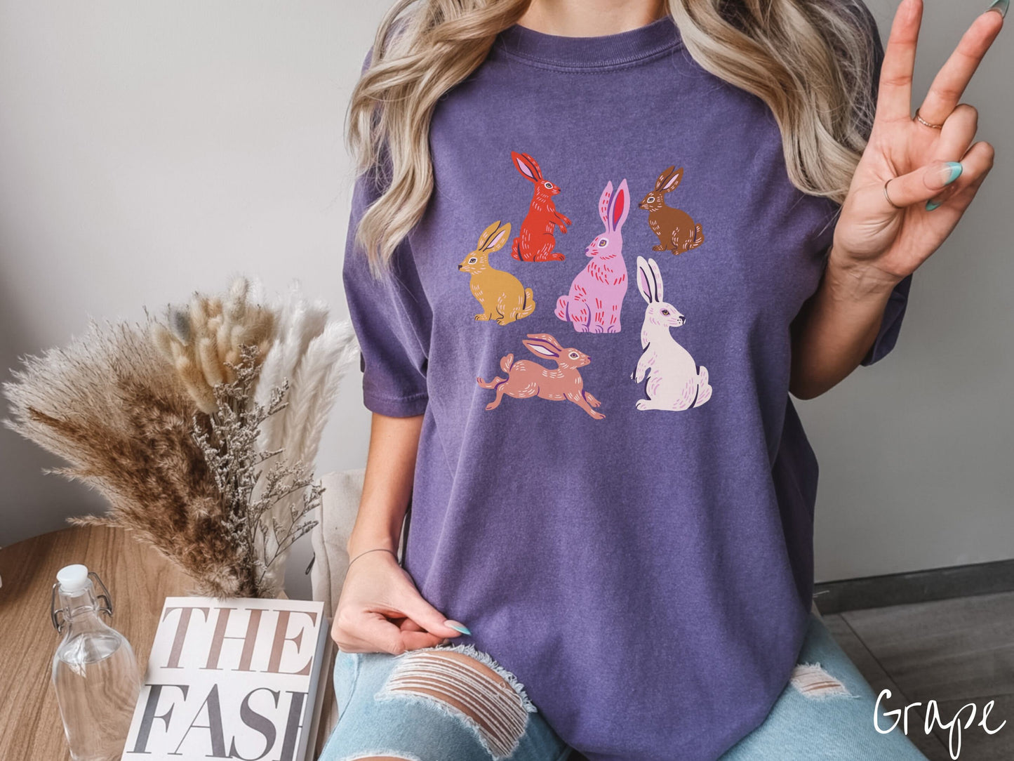 A woman wearing a cute, vintage grape colored shirt with six colored bunny rabbits in different stances and postures. One is red, another brown, yellow, pink, light brown, and white.