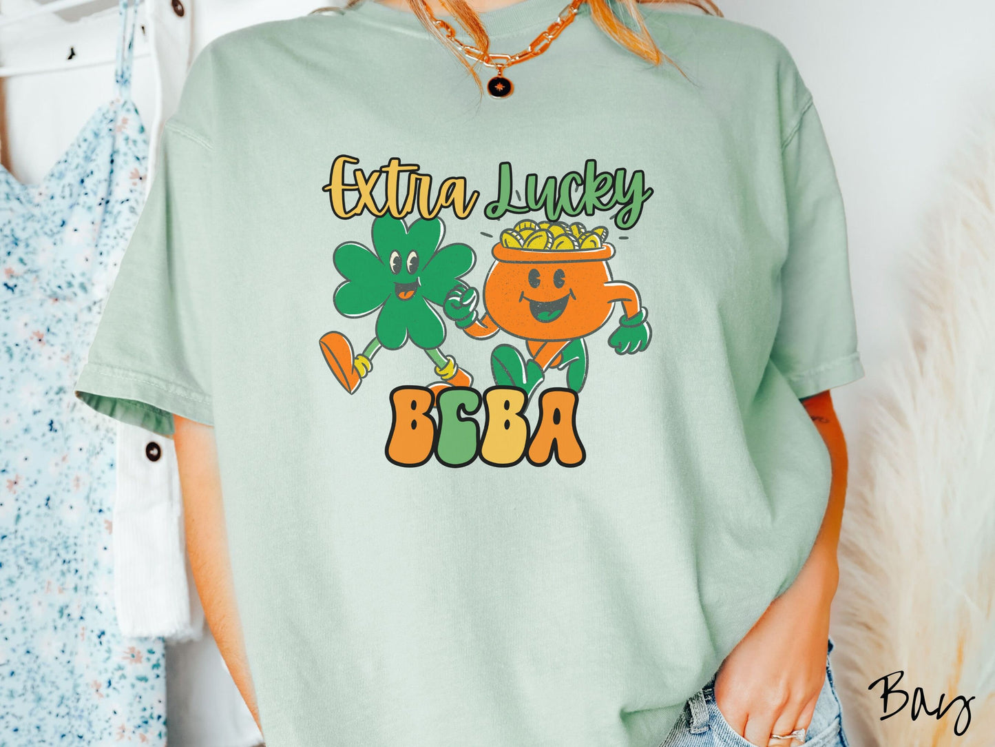 A woman wearing a vintage, bay colored shirt with the text Extra Lucky BCBA in yellow, orange, and green font. Between the text are a green clover and orange pot of gold smiling and walking together in step.