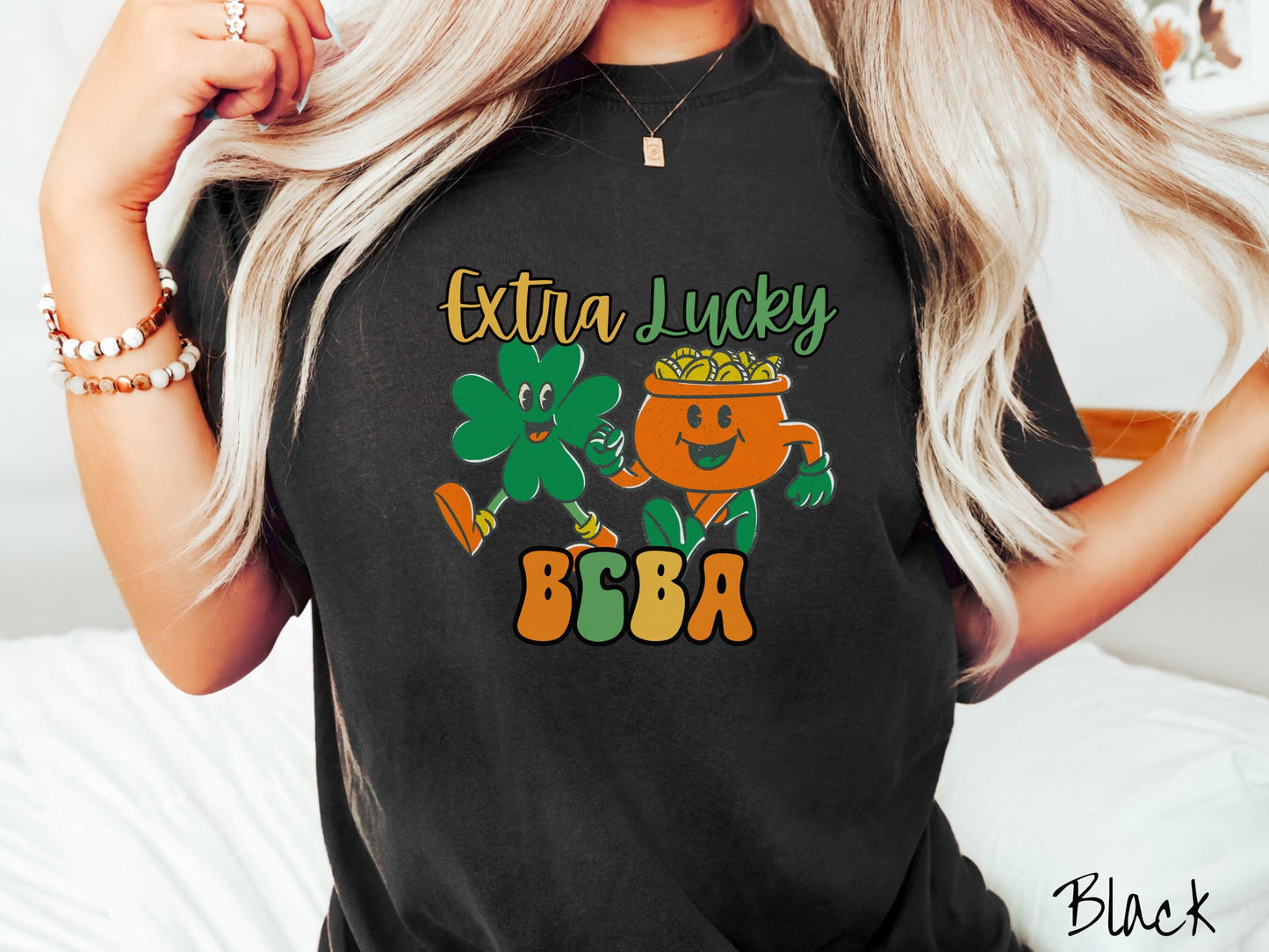 A woman wearing a vintage, black colored shirt with the text Extra Lucky BCBA in yellow, orange, and green font. Between the text are a green clover and orange pot of gold smiling and walking together in step.