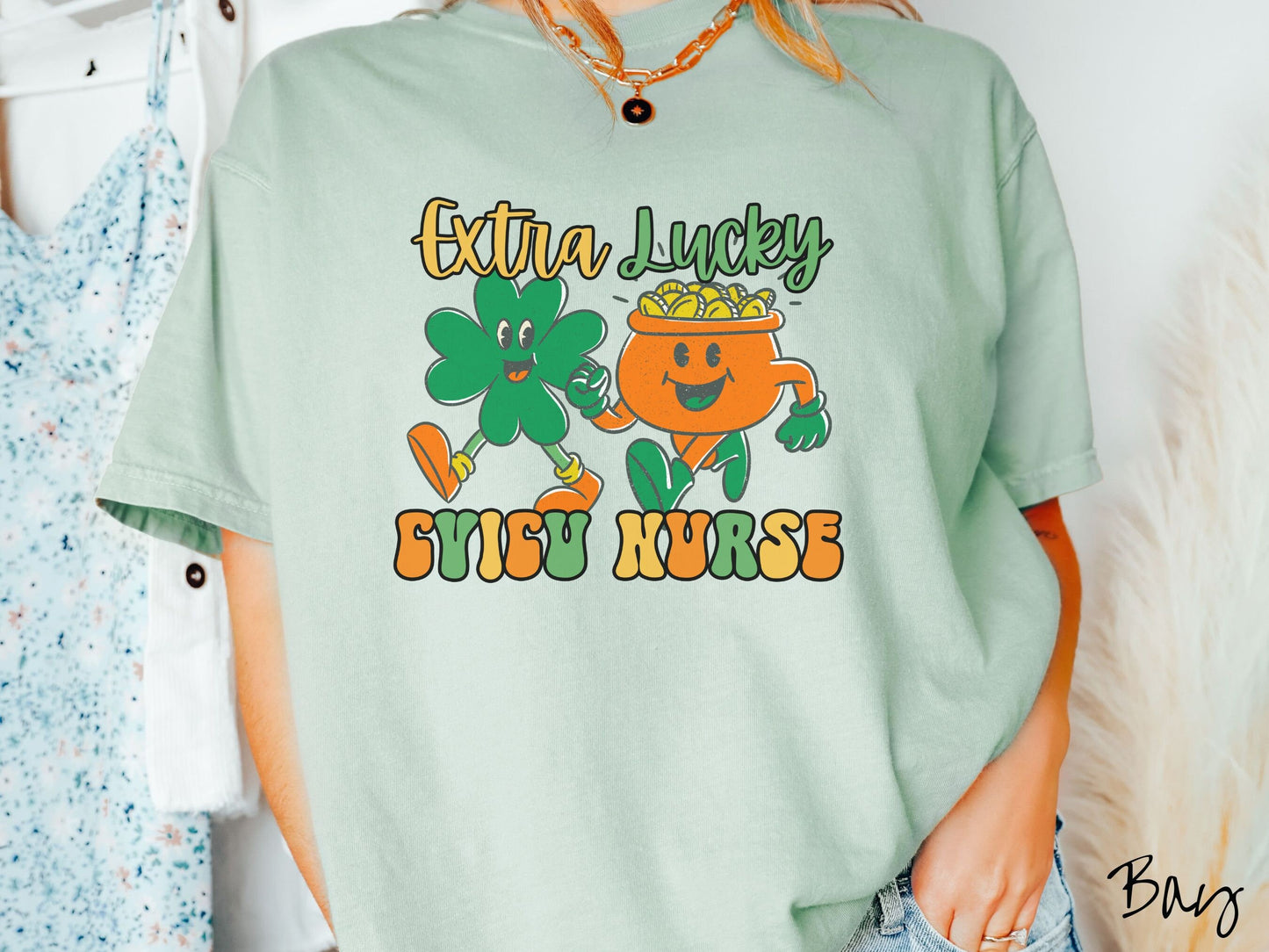 A woman wearing a vintage, bay colored shirt with the text Extra Lucky CVICU Nurse in yellow, orange, and green font. Between the text are a green clover and orange pot of gold smiling and walking together in step.