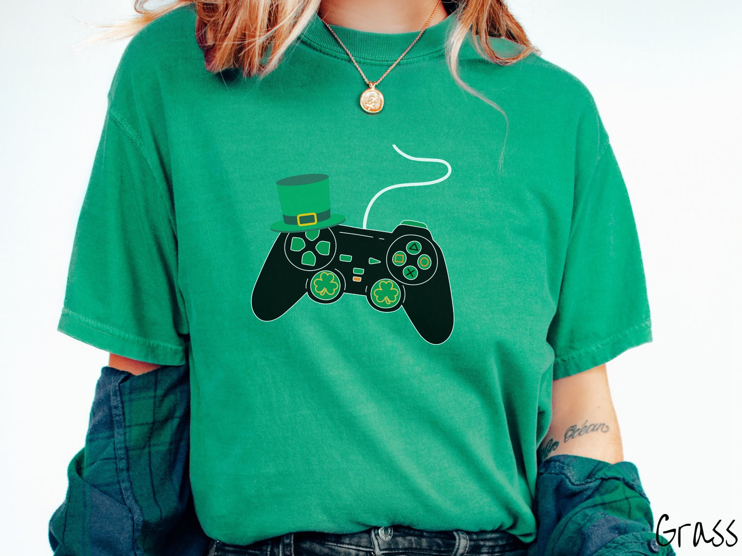 A woman wearing a vintage, grass colored shirt with a black video game controller with green buttons and joysticks, the analog control sticks are in the shape of green leaf clovers, and there is a green top hat hanging off the top left trigger.