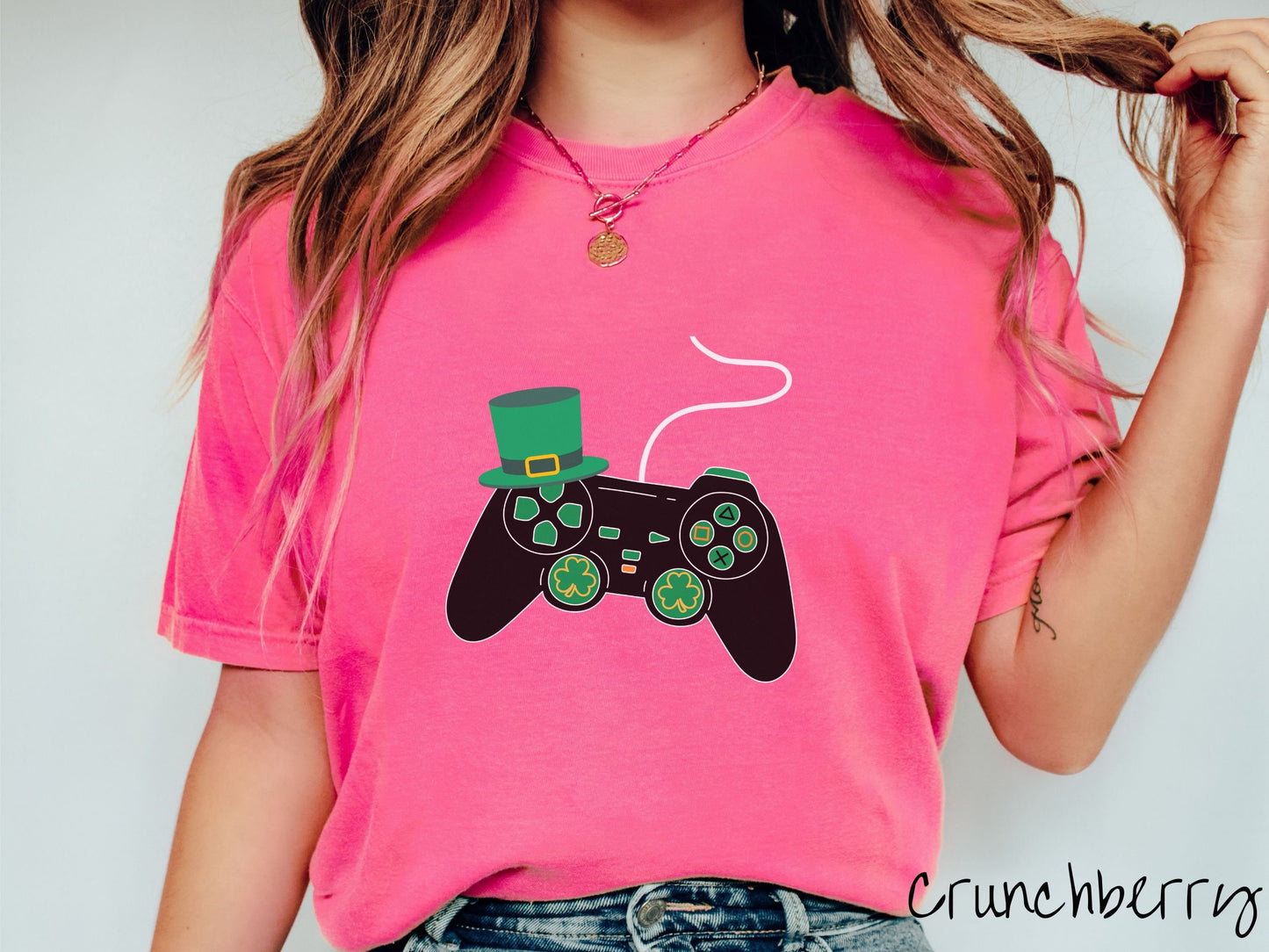 A woman wearing a vintage, crunchberry colored shirt with a black video game controller with green buttons and joysticks, the analog control sticks are in the shape of green leaf clovers, and there is a green top hat hanging off the top left trigger.