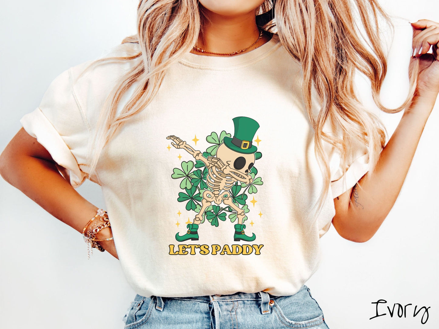 A woman wearing a vintage, ivory colored shirt with the text Lets Paddy and below that is a leprechaun wearing a green top hat and green elf shoes doing the dab dance and there are green clovers and golden stars in the background.