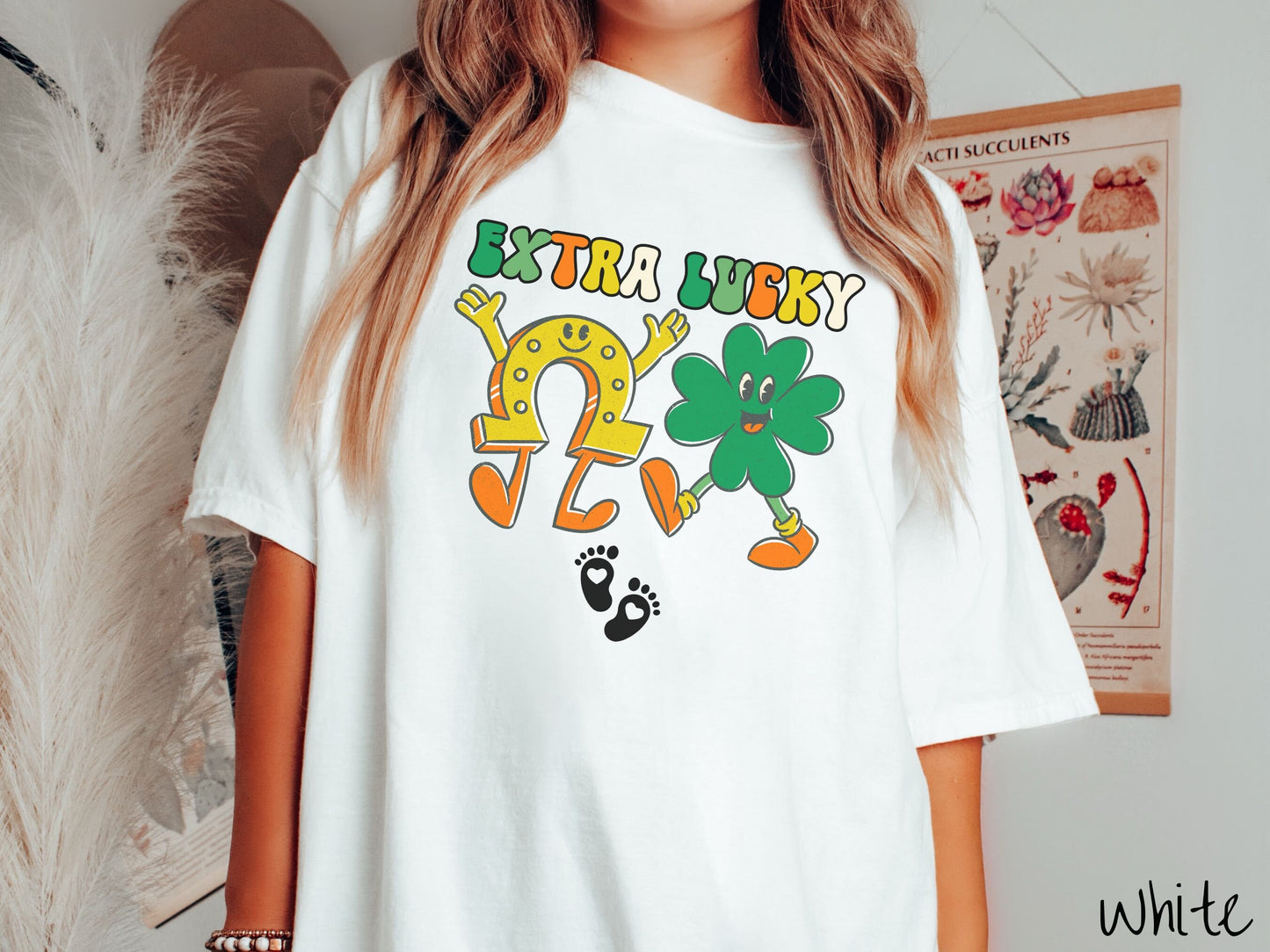 A woman wearing a vintage, white colored shirt with text Extra Lucky in green, white, and orange font. Below are a yellow horseshoe with a smile and its hands up, a green clover smiling, and two white baby footprints with hearts.