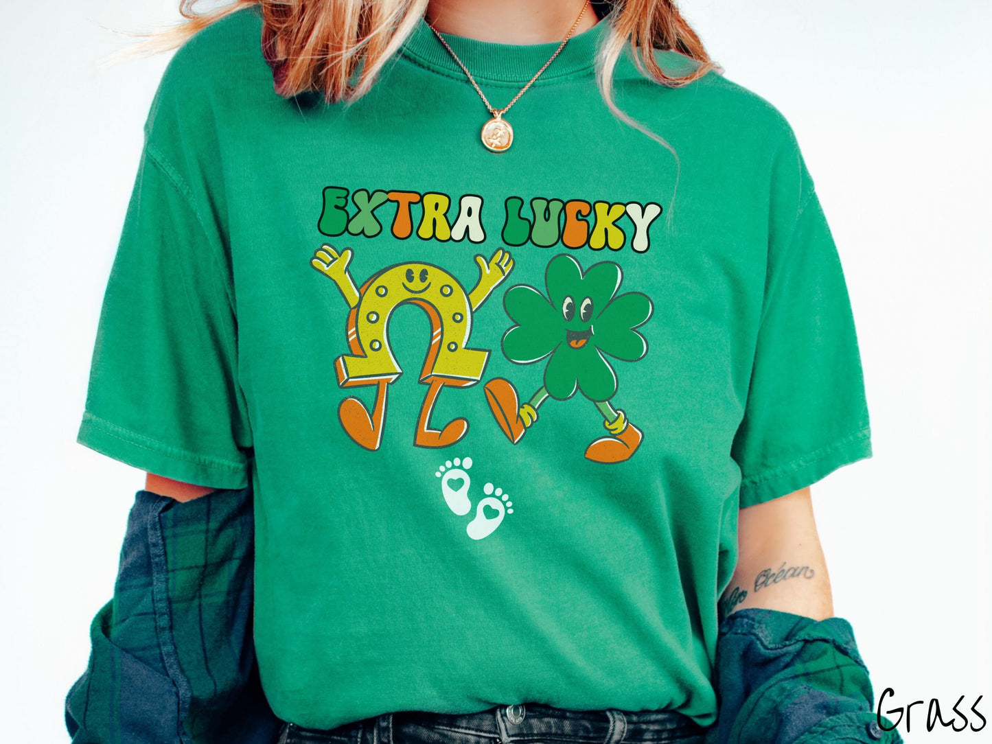 A woman wearing a vintage, grass colored shirt with text Extra Lucky in green, white, and orange font. Below are a yellow horseshoe with a smile and its hands up, a green clover smiling, and two white baby footprints with hearts.