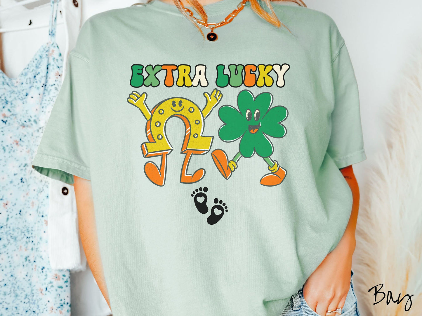 A woman wearing a vintage, bay colored shirt with text Extra Lucky in green, white, and orange font. Below are a yellow horseshoe with a smile and its hands up, a green clover smiling, and two white baby footprints with hearts.