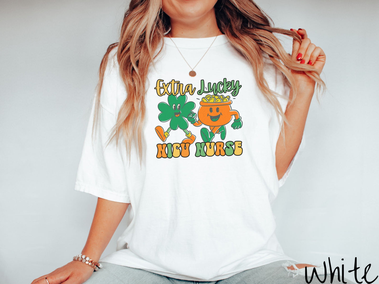 A woman wearing a vintage, white colored shirt with the text Extra Lucky NICU Nurse in yellow, orange, and green font. Between the text are a green clover and orange pot of gold smiling and walking together in step.