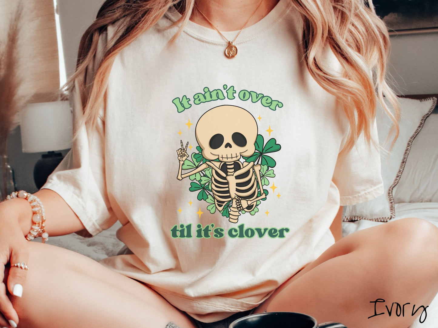 A woman wearing a vintage, ivory colored shirt with the text It aint over til its clover and in between the text is a smiling skeleton holding a peace sign with its right hand, with many green three leaf clovers and golden stars in the background.