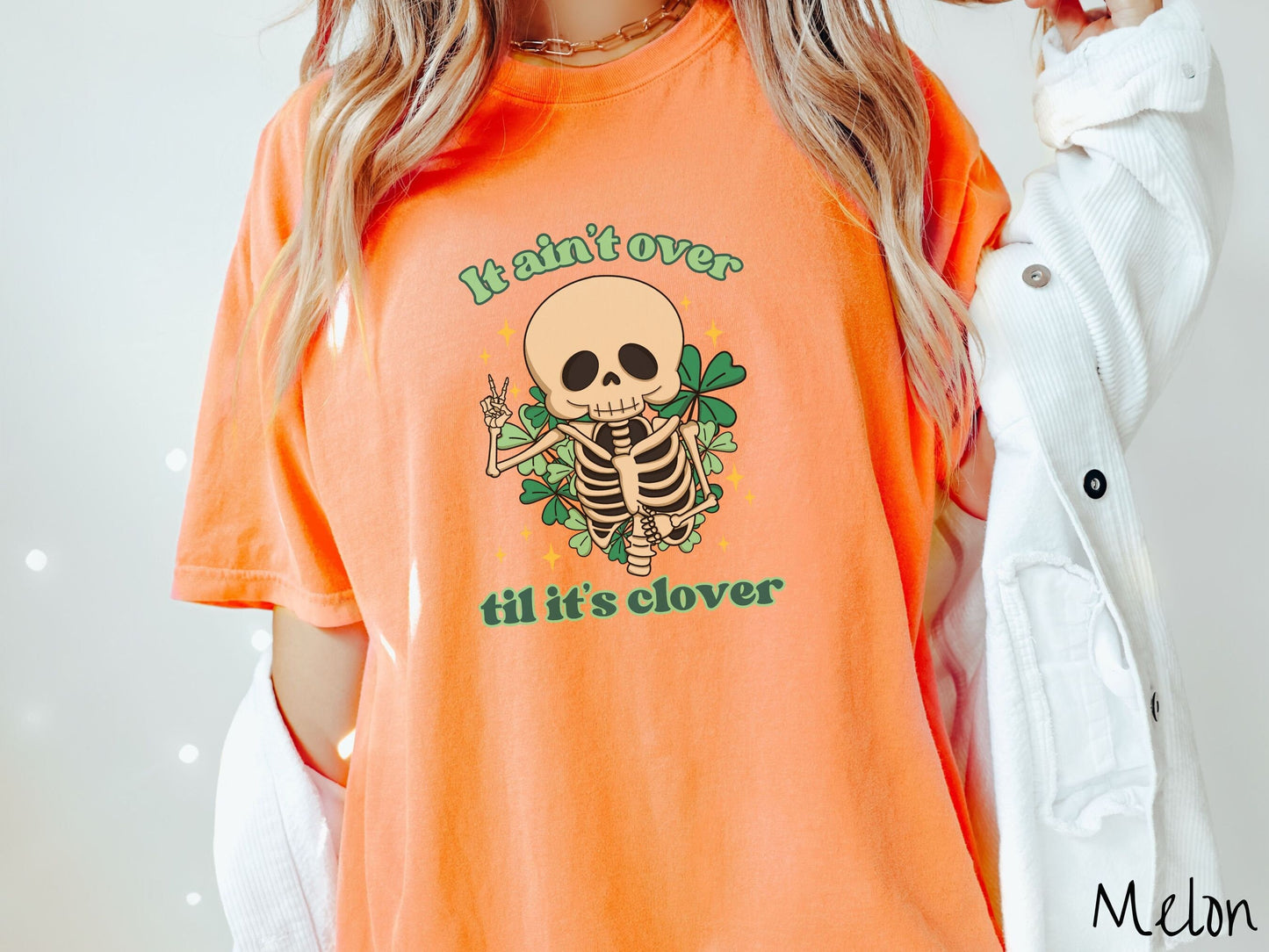 A woman wearing a vintage, melon colored shirt with the text It aint over til its clover and in between the text is a smiling skeleton holding a peace sign with its right hand, with many green three leaf clovers and golden stars in the background.
