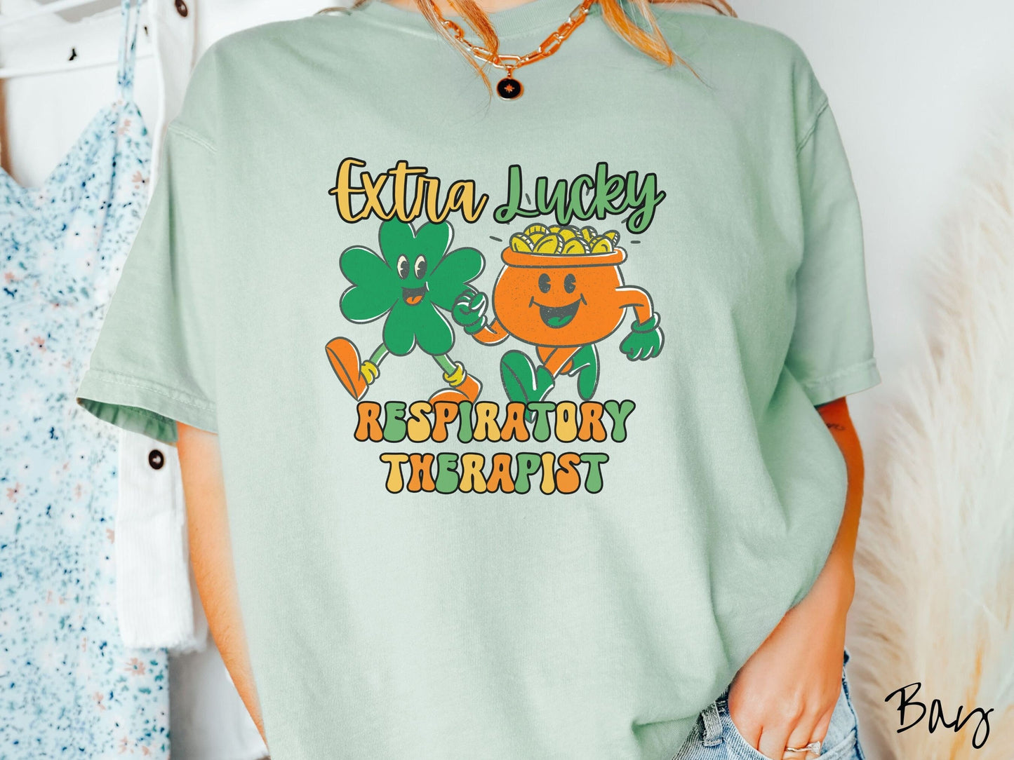 A woman wearing a vintage, bay colored shirt with the text Extra Lucky Respiratory Therapist in yellow, orange, and green font. Between the text are a green clover and orange pot of gold smiling and walking together in step.