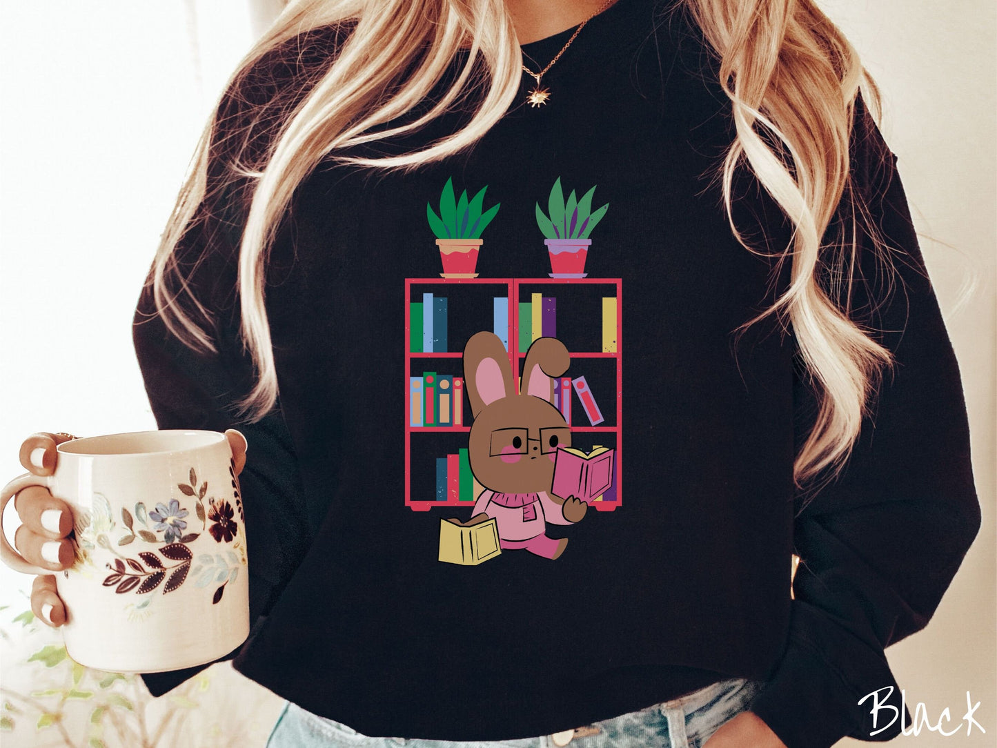 A woman wearing a cute, vintage black colored sweatshirt with a pink-cheeked, brown bunny rabbit wearing glasses reading a pink book in one hand, and a yellow book in the other. In the background is a bookshelf with green plants on top.
