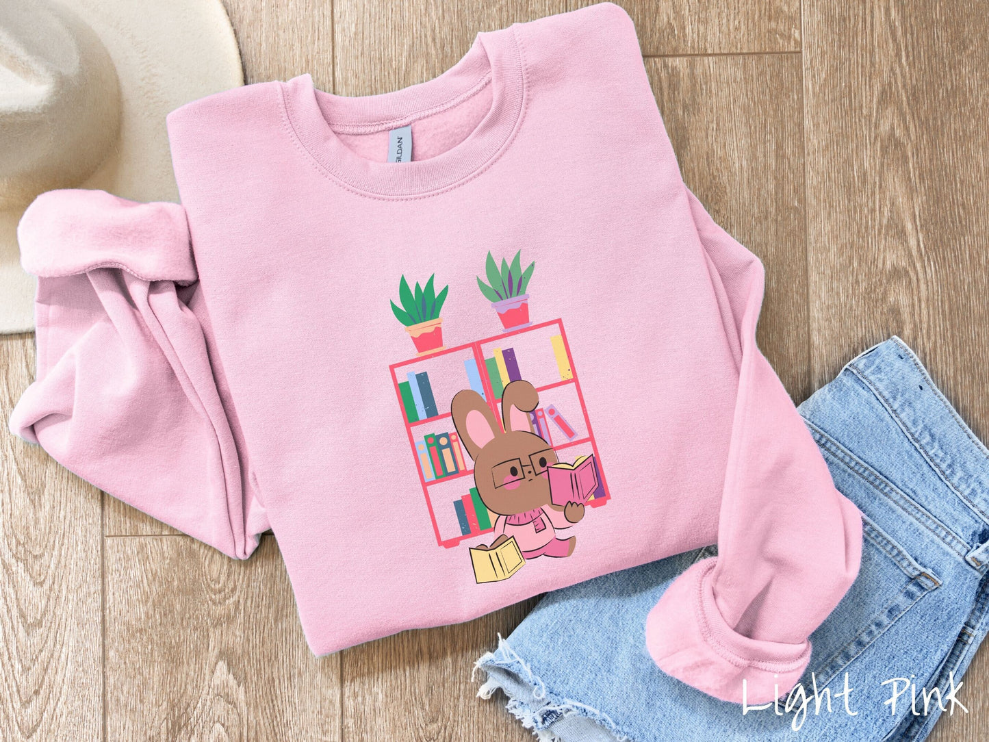 A cute, vintage light pink colored sweatshirt with a pink-cheeked, brown bunny rabbit wearing glasses reading a pink book in one hand, and a yellow book in the other. In the background is a bookshelf with green plants on top.