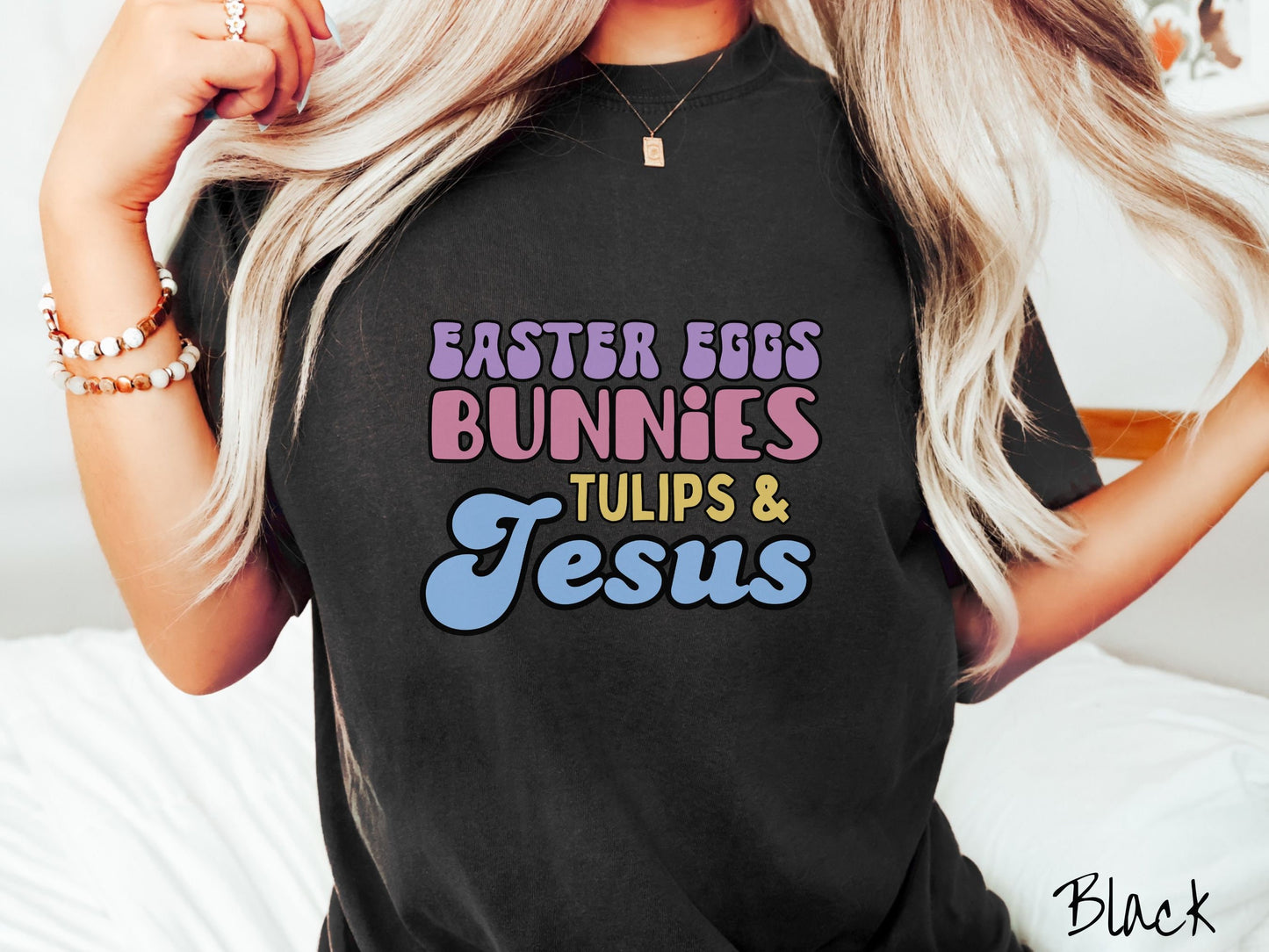 A woman wearing a cute, vintage black colored shirt with text Easter Eggs, Bunnies, Tulips, & Jesus in purple, pink, yellow, and blue font.