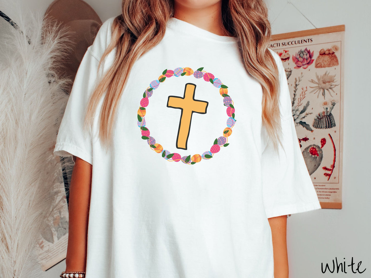 A woman wearing a cute, vintage white colored shirt with a golden cross with black outline in the center surrounded by a wreath of colorful Easter eggs forming a circle around the cross, intertwined with green leaves.