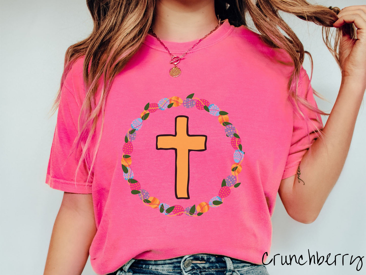 A woman wearing a cute, vintage crunchberry colored shirt with a golden cross with black outline in the center surrounded by a wreath of colorful Easter eggs forming a circle around the cross, intertwined with green leaves.
