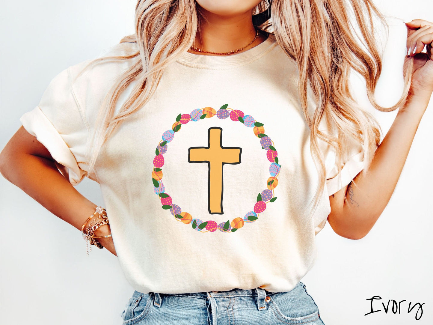 A woman wearing a cute, vintage ivory colored shirt with a golden cross with black outline in the center surrounded by a wreath of colorful Easter eggs forming a circle around the cross, intertwined with green leaves.