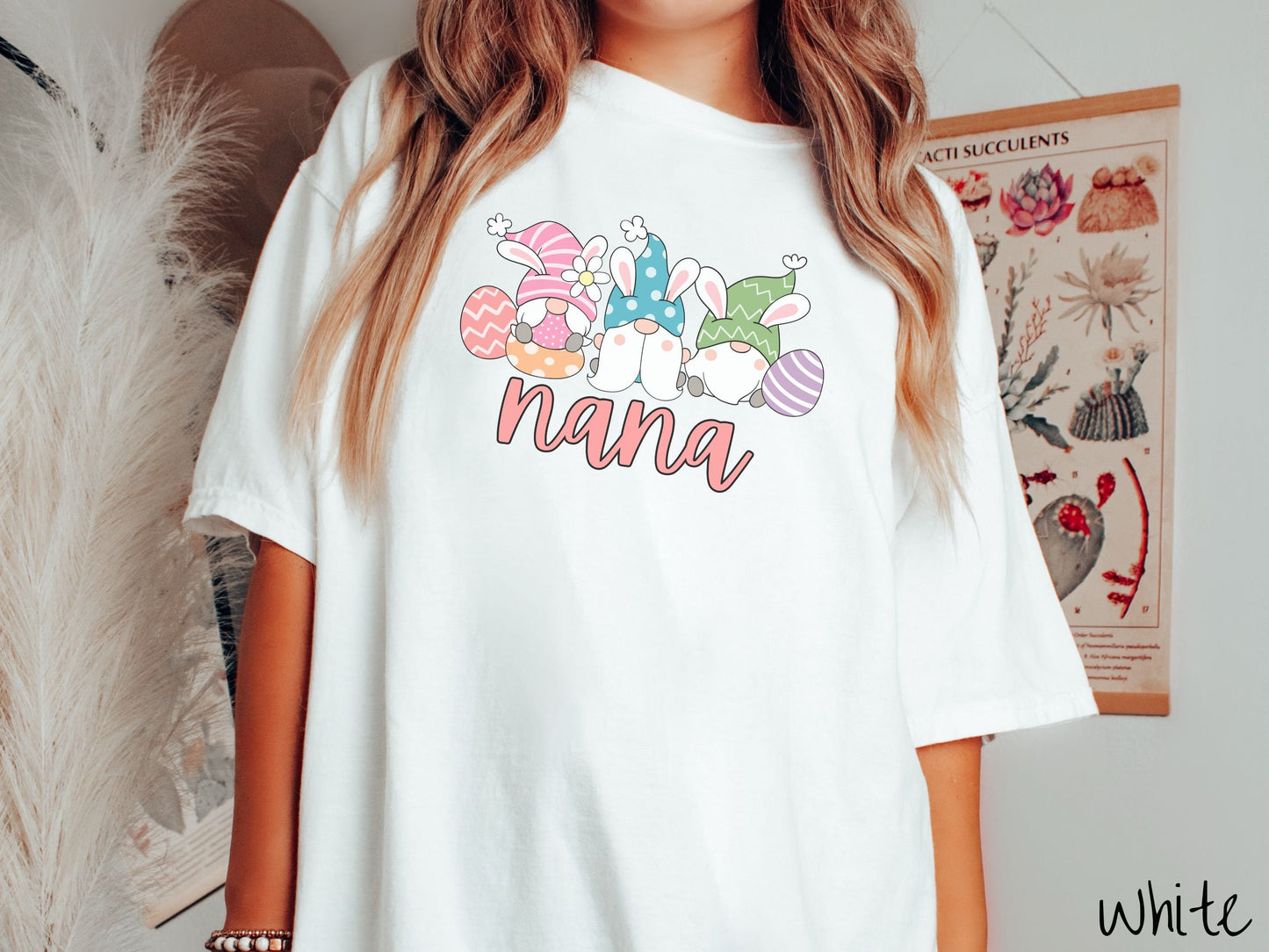 A woman wearing a cute, vintage white colored shirt with text Nana below three gnomes with rabbit ears sitting next to each other. The left one is wearing pink, the middle blue, and right in green, with colorful easter eggs scattered about.