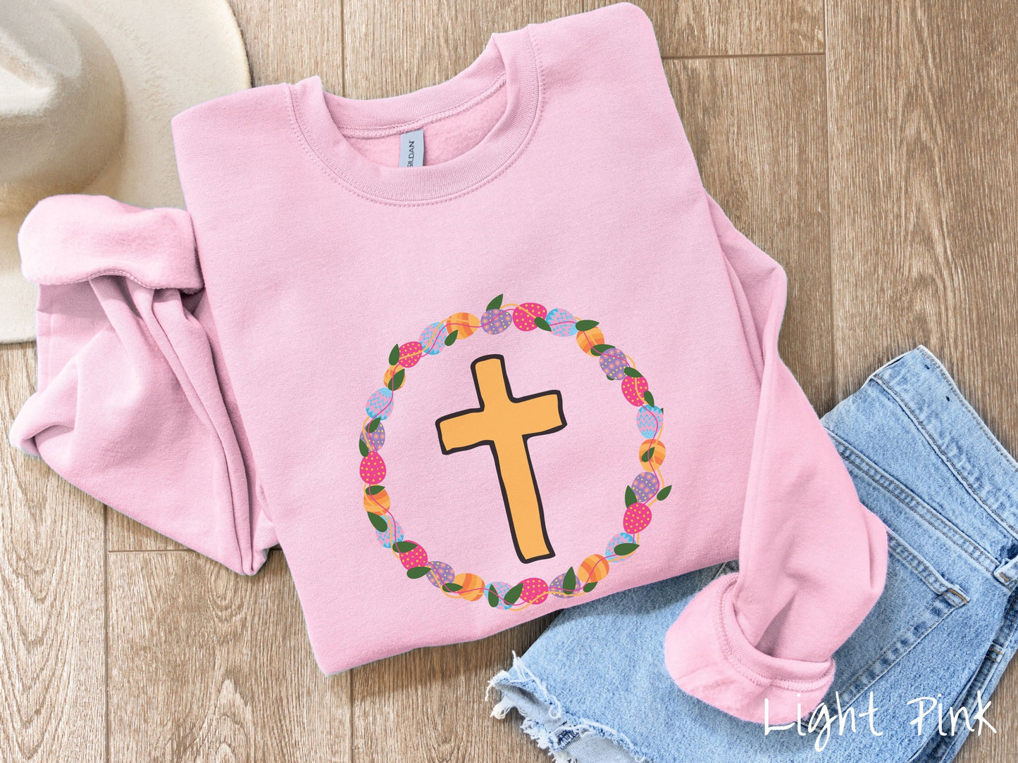 A cute, vintage light pink colored sweatshirt with a golden cross with black outline in the center surrounded by a wreath of colorful Easter eggs forming a circle around the cross, intertwined with green leaves.
