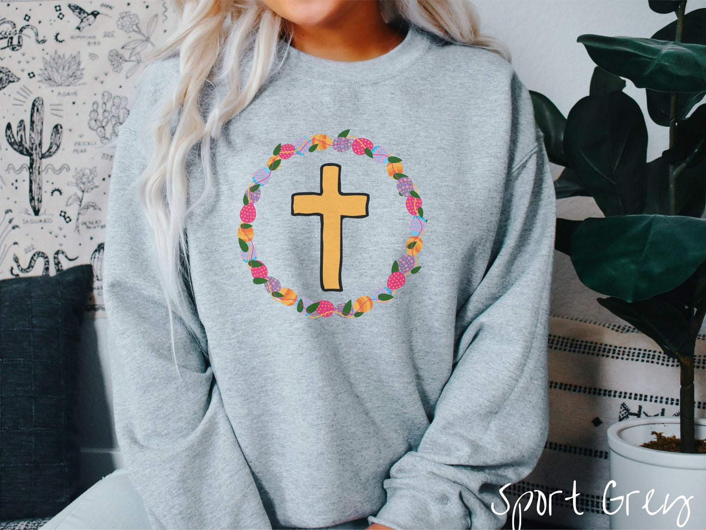 A woman wearing a cute, vintage sport grey colored sweatshirt with a golden cross with black outline in the center surrounded by a wreath of colorful Easter eggs forming a circle around the cross, intertwined with green leaves.