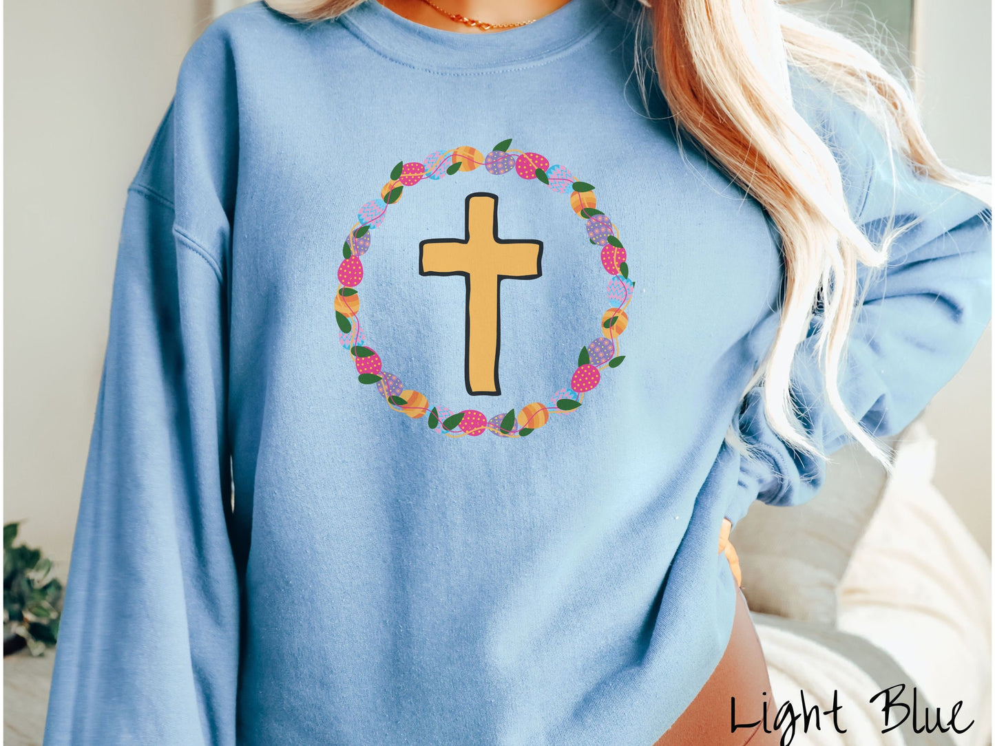 A woman wearing a cute, vintage light blue colored sweatshirt with a golden cross with black outline in the center surrounded by a wreath of colorful Easter eggs forming a circle around the cross, intertwined with green leaves.