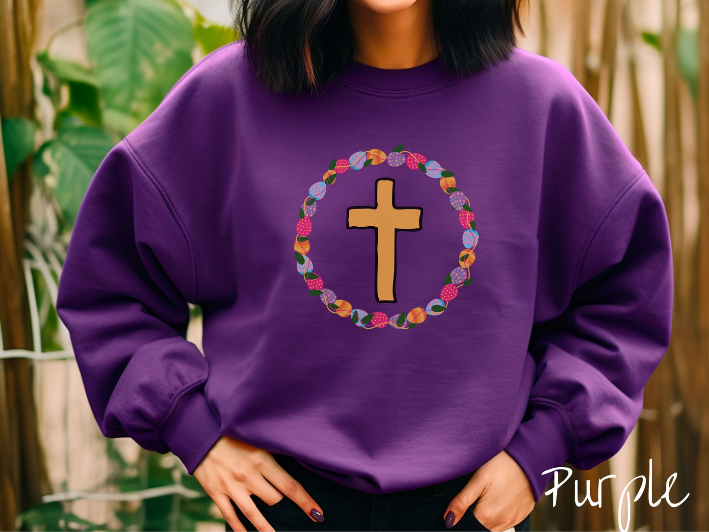 A woman wearing a cute, vintage purple colored sweatshirt with a golden cross with black outline in the center surrounded by a wreath of colorful Easter eggs forming a circle around the cross, intertwined with green leaves.