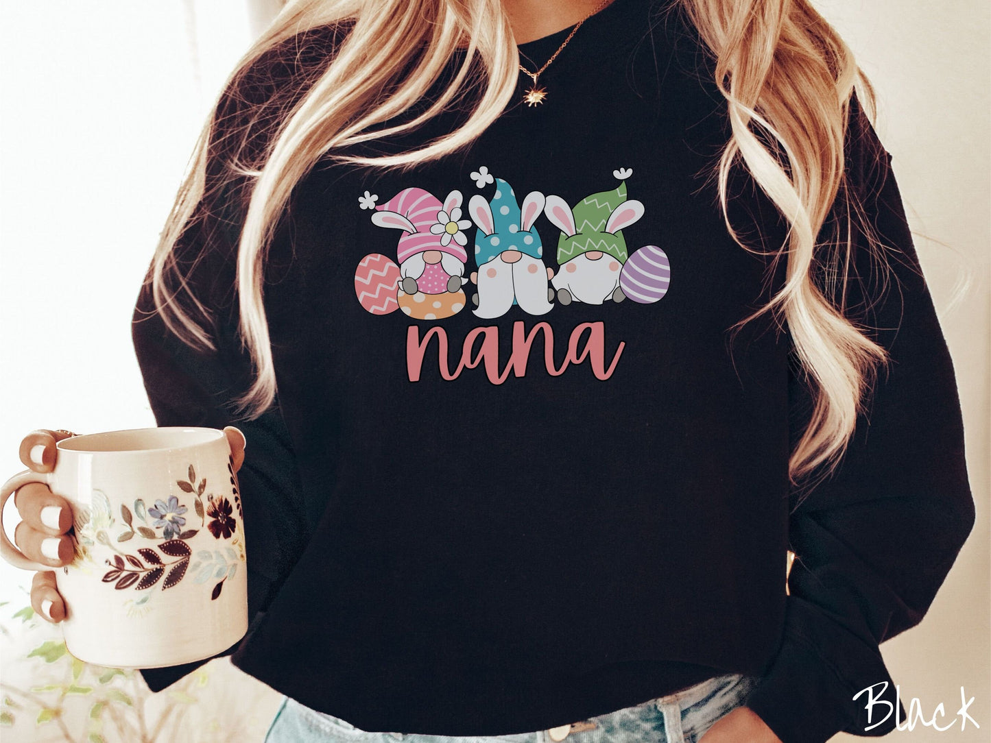 A woman wearing a cute, vintage black colored sweatshirt with the text Nana below three gnomes with rabbit ears sitting next to each other. The left one is wearing pink, the middle blue, and right in green, with colorful easter eggs scattered about.