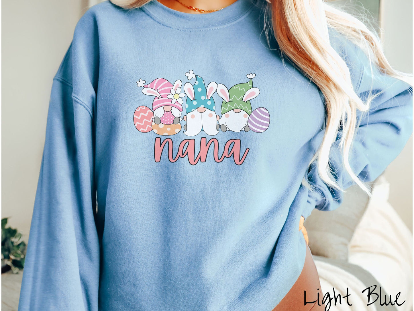 A woman wearing a cute, vintage light blue colored sweatshirt with text Nana below three gnomes with rabbit ears sitting next to each other. The left one is wearing pink, the middle blue, and right in green, with colorful easter eggs scattered about.