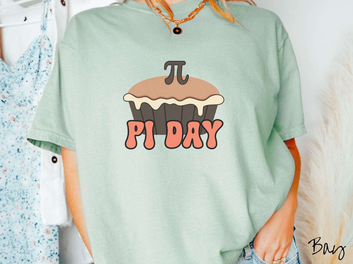 A woman wearing a cute, vintage bay colored comfort colors shirt with the text Pi Day in orange font, above that is a brown pumpkin pie with a pi symbol on the top of the pie.