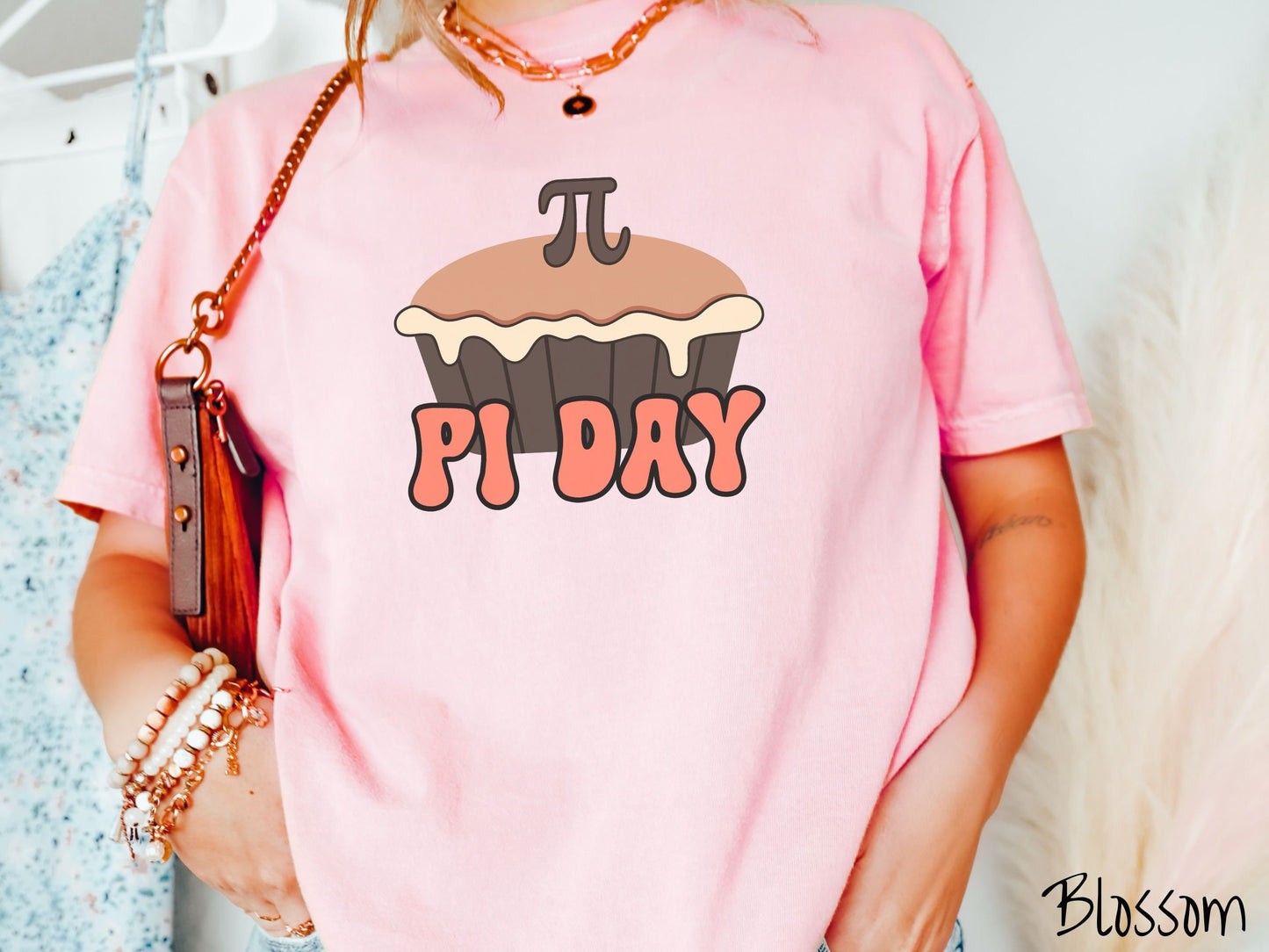 A woman wearing a cute, vintage blossom colored comfort colors shirt with the text Pi Day in orange font, above that is a brown pumpkin pie with a pi symbol on the top of the pie.