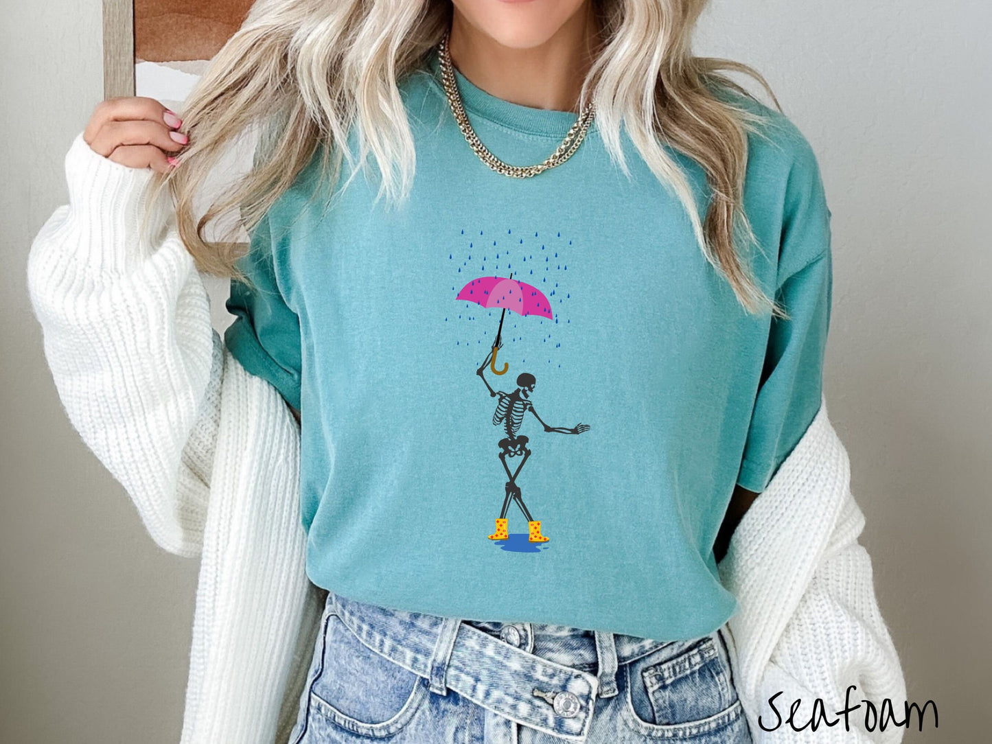 A woman wearing a cute seafoam teal colored shirt with a dark grey female skeleton dancing in the rain with a pink and purple umbrella and wearing yellow with red polka dot rain boots.
