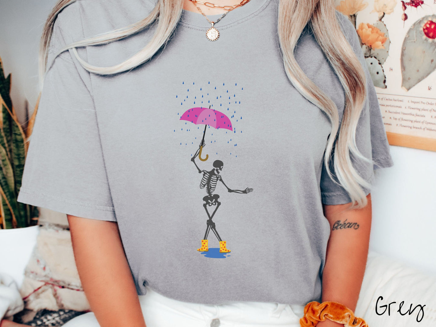 A woman wearing a cute grey colored shirt with a dark grey female skeleton dancing in the rain with a pink and purple umbrella and wearing yellow with red polka dot rain boots.