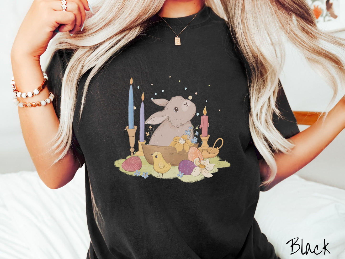 A woman wearing a cute, vintage black colored shirt with a gray bunny rabbit sitting in between three lit candles, some red, orange, and purple eggs, and yellow and white flowers.