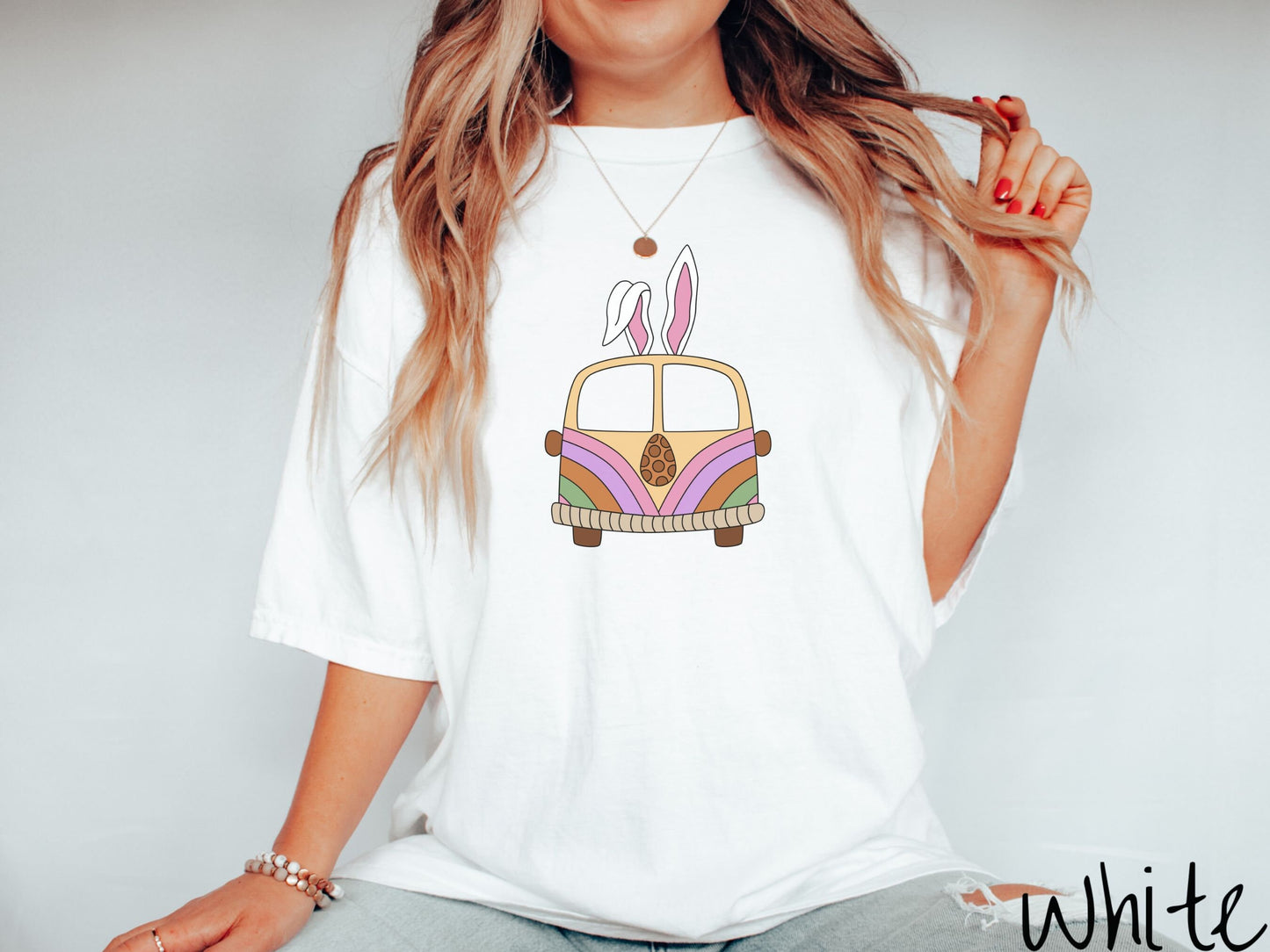 A woman wearing a cute, vintage white colored shirt with an old-fashioned, hippie style van with pastel-colored stripes on the front and light orange paint covering the rest. The van has a brown egg on the front and two, floppy bunny ears on top.
