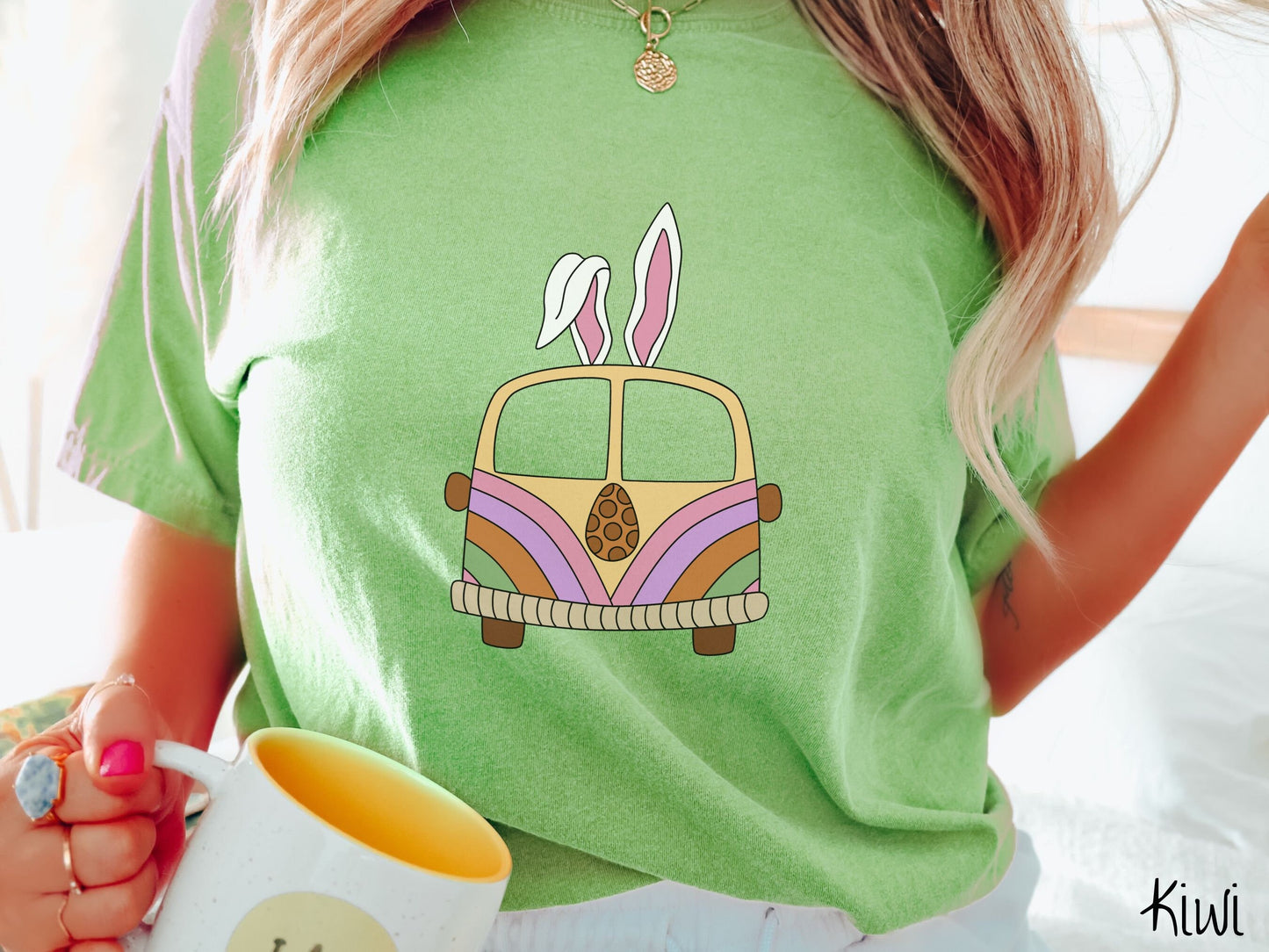 A woman wearing a cute, vintage kiwi colored shirt with an old-fashioned, hippie style van with pastel-colored stripes on the front and light orange paint covering the rest. The van has a brown egg on the front and two, floppy bunny ears on top.
