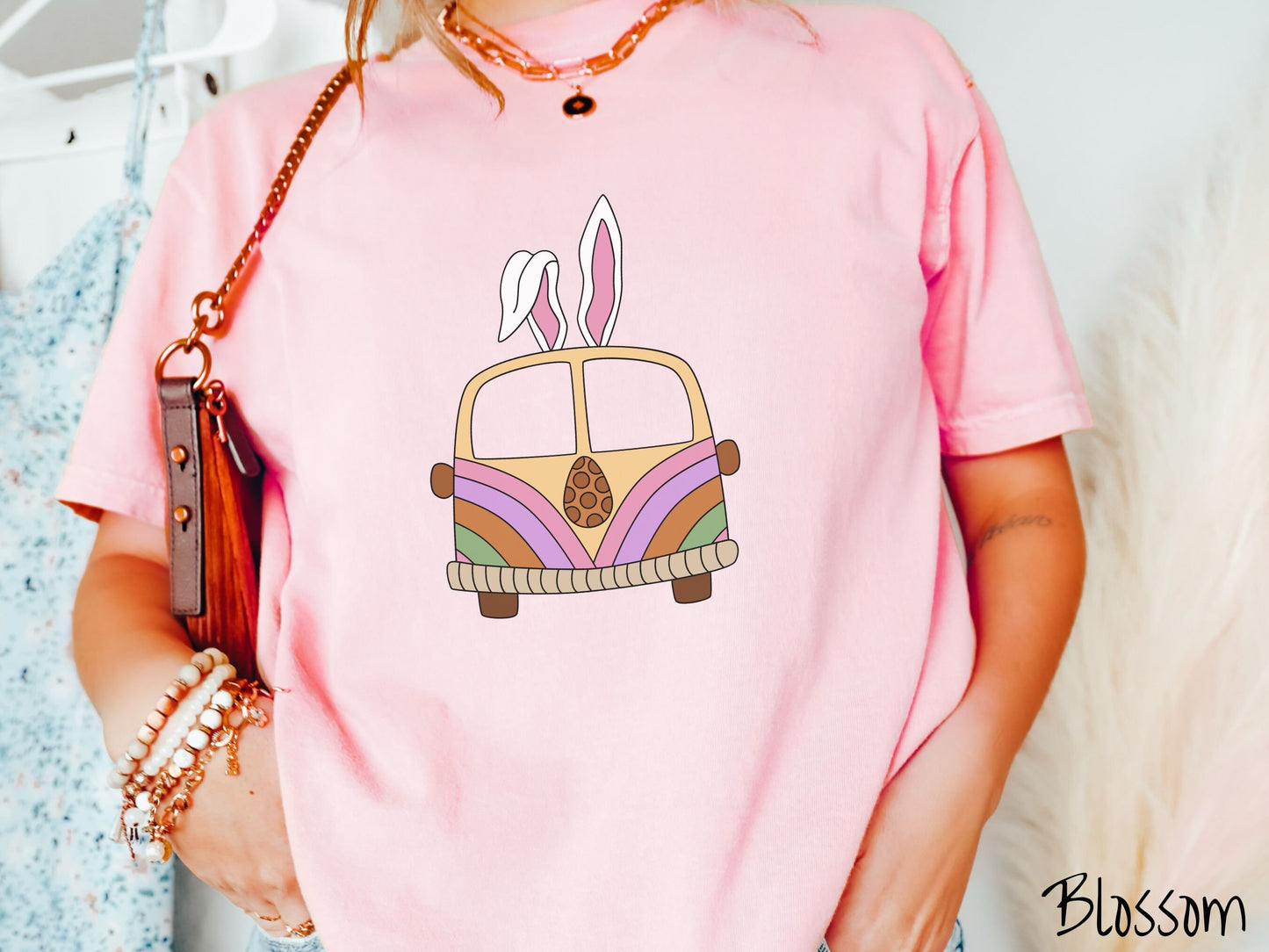 A woman wearing a cute, vintage blossom colored shirt with an old-fashioned, hippie style van with pastel-colored stripes on the front and light orange paint covering the rest. The van has a brown egg on the front and two, floppy bunny ears on top.