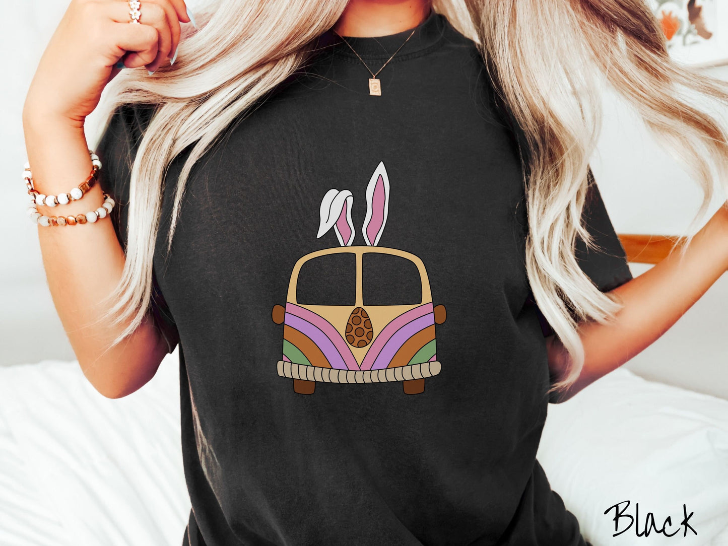 A woman wearing a cute, vintage black colored shirt with an old-fashioned, hippie style van with pastel-colored stripes on the front and light orange paint covering the rest. The van has a brown egg on the front and two, floppy bunny ears on top.