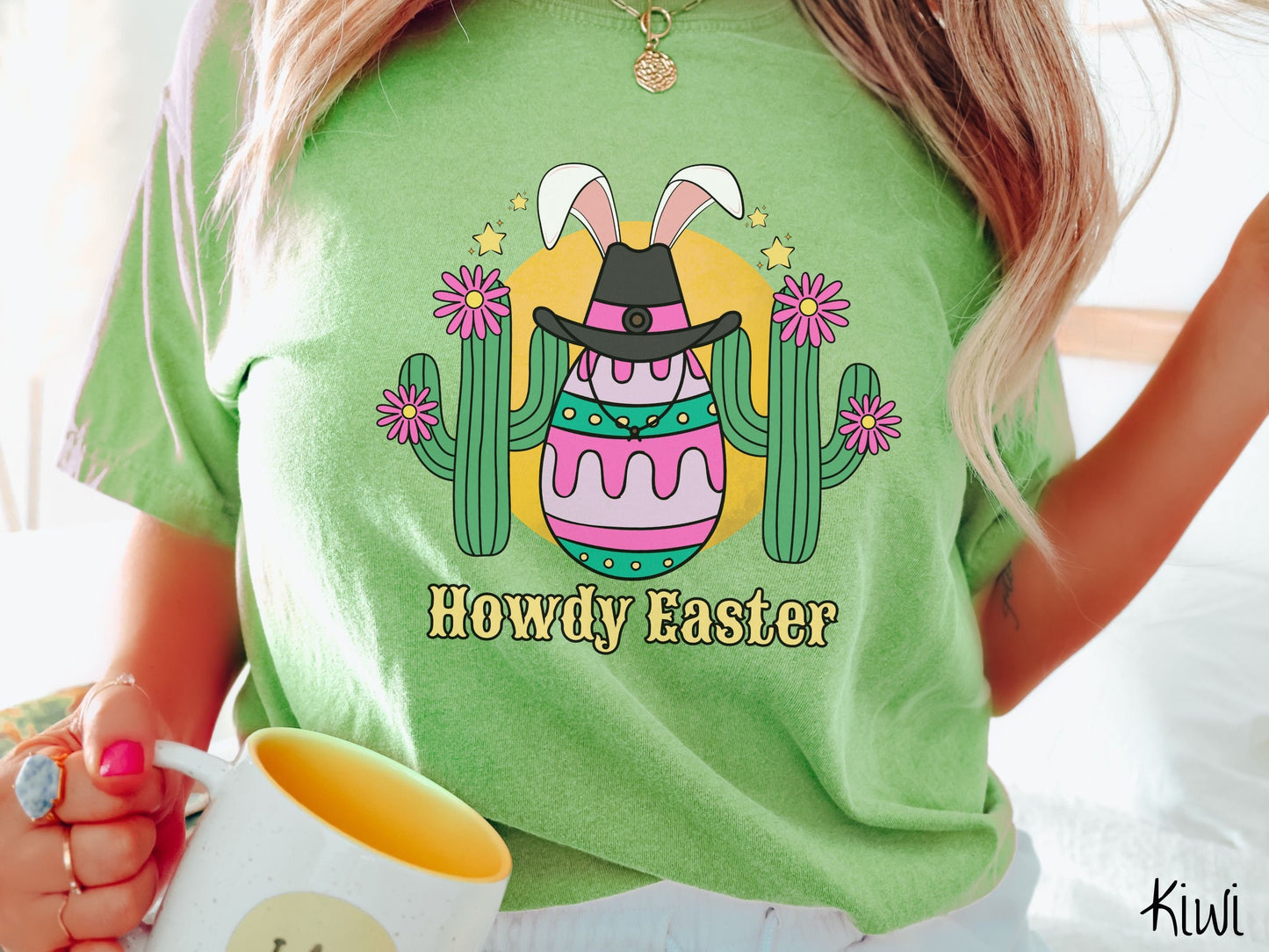 A woman wearing a cute, vintage kiwi colored shirt with an Easter egg with green, pink and purple stripes, wearing a black cowboy hat and white bunny ears, and it’s standing in between two green cacti with pink flowers in front of the sun.