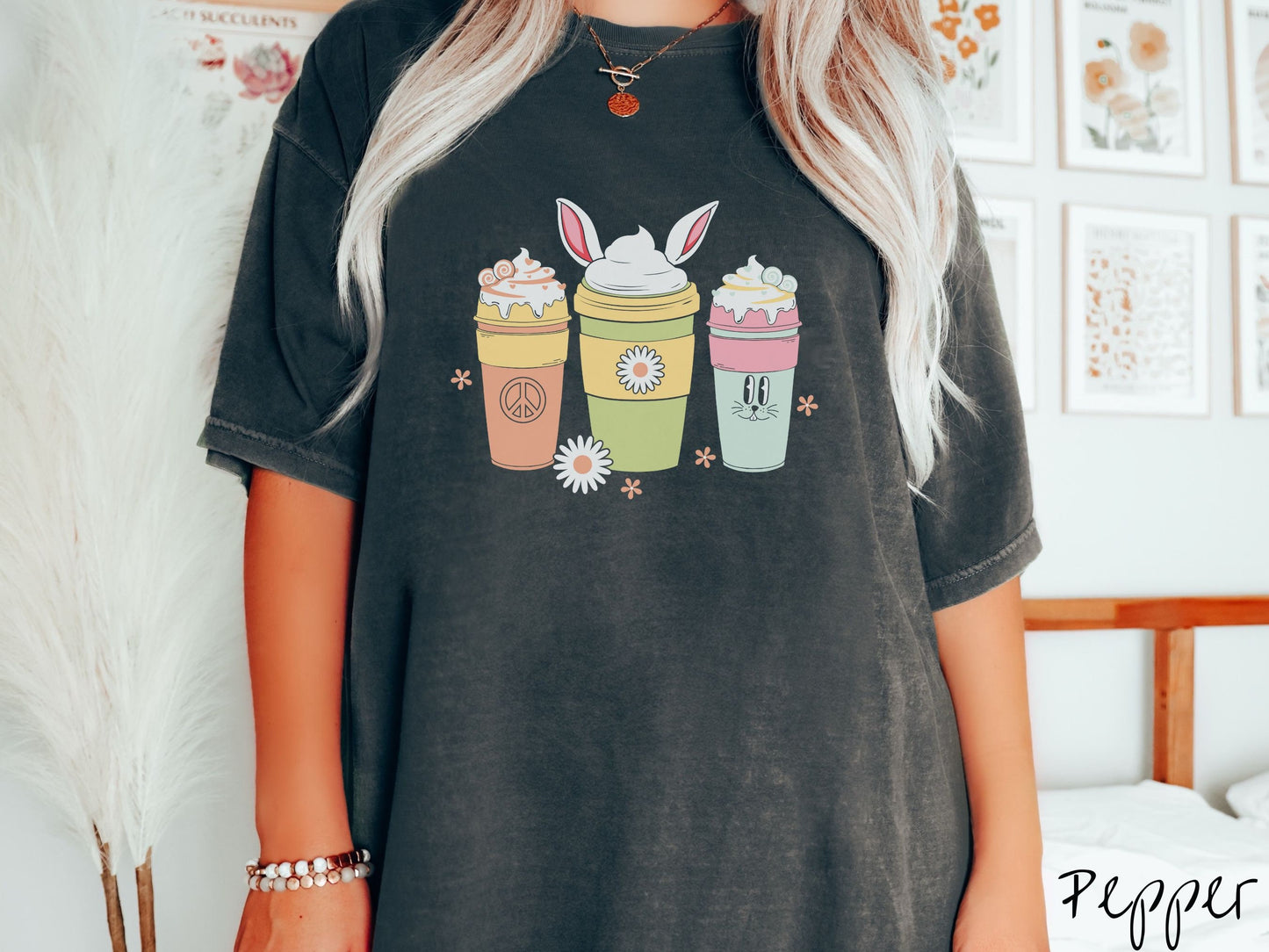 A woman wearing a cute, vintage pepper colored shirt with three coffee cups on the front. One is orange with whipped cream and a peace sign. The middle is green with whipped cream, and bunny ears, and the third is light blue with whipped cream.