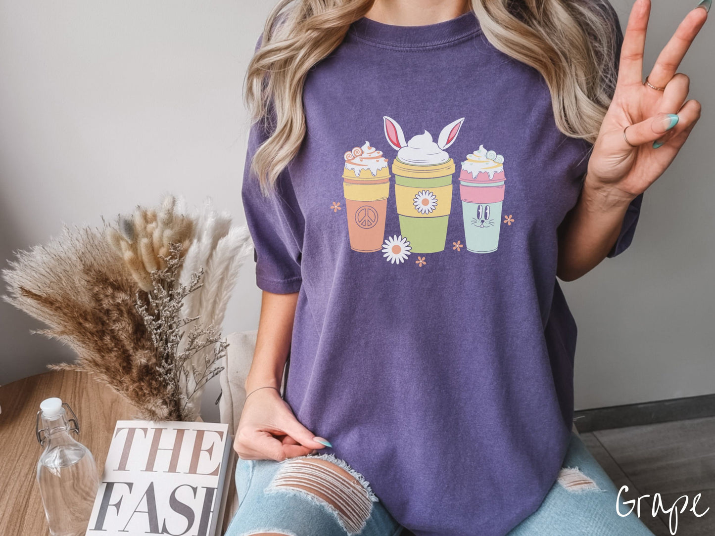 A woman wearing a cute, vintage grape colored shirt with three coffee cups on the front. One is orange with whipped cream and a peace sign. The middle is green with whipped cream, and bunny ears, and the third is light blue with whipped cream.